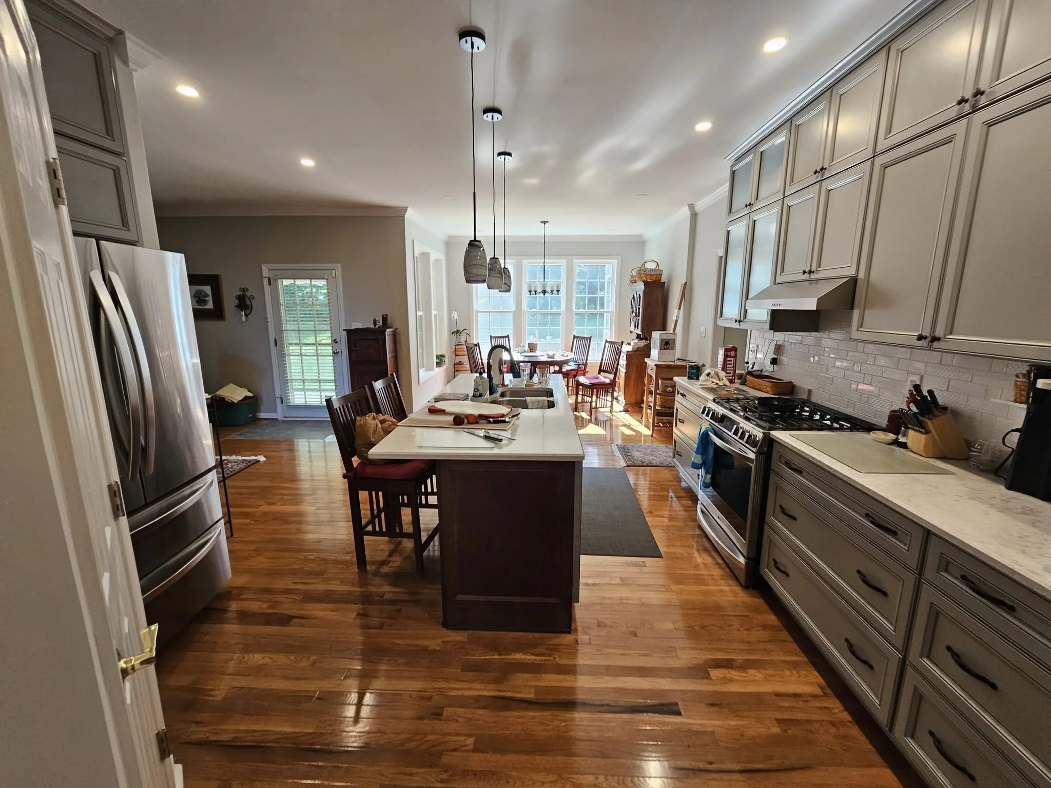 A spacious kitchen with wooden floors, gray cabinets, a large kitchen island with a white countertop, and stainless steel appliances including a refrigerator and oven. There are pendant lights above the island and a dining area with a table and chair