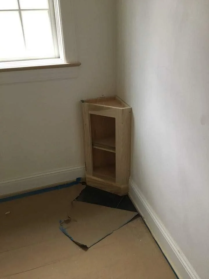 Small unfinished wooden cabinet placed in the corner of a room, next to a window, on tape and cardboard covering the floor.