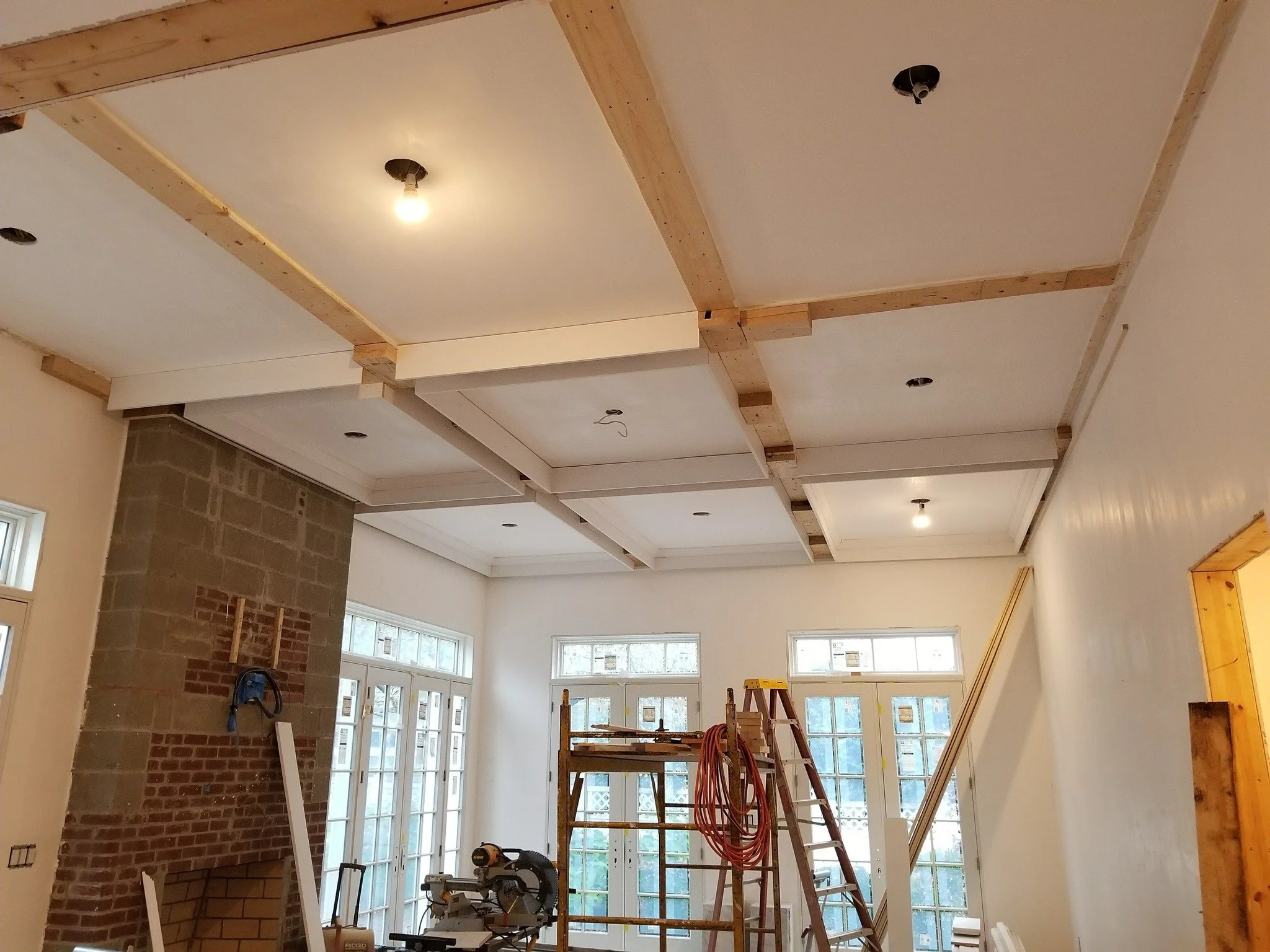 Interior of a house under construction with drywall, ceiling lights, a brick fireplace, and construction tools and scaffolding inside.