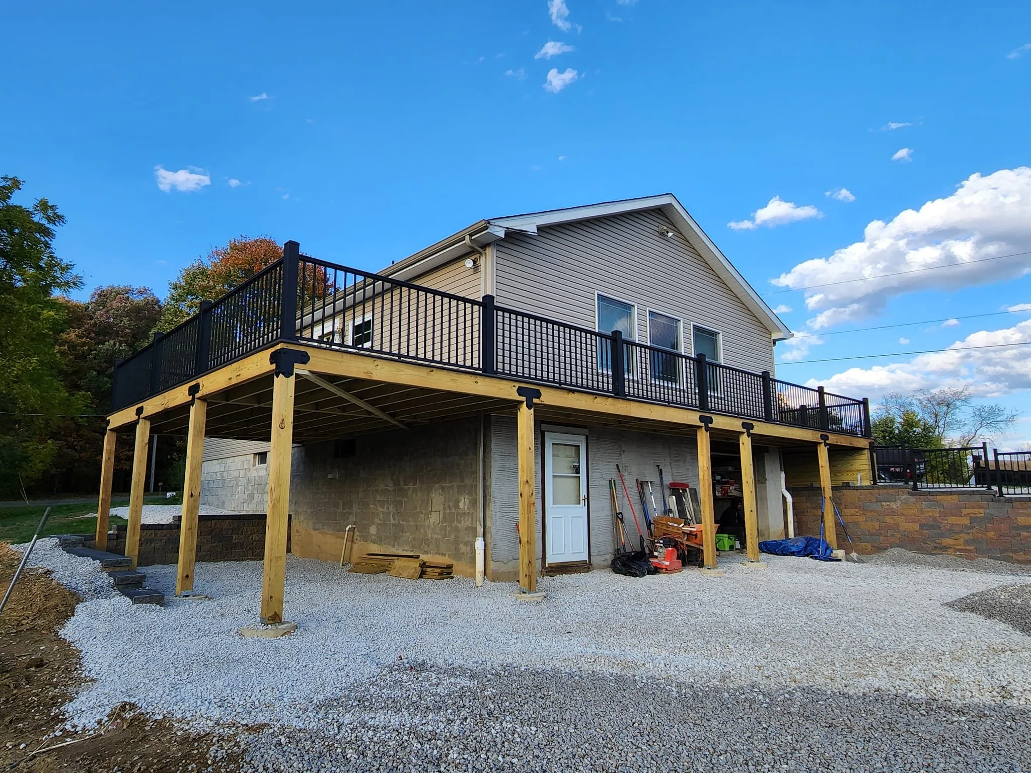 Two-story house with a balcony under construction, elevated on wooden posts, with a gravel ground and construction tools nearby, under a partly cloudy sky.