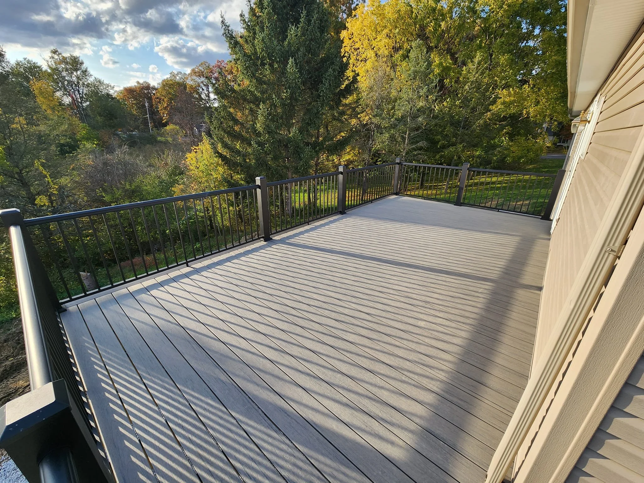 Empty wooden deck with black metal railing overlooking trees and a partly cloudy sky.