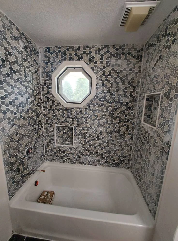 Bathroom shower with hexagonal tile walls, small built-in wall niches, small octagonal window, and a white shower pan.