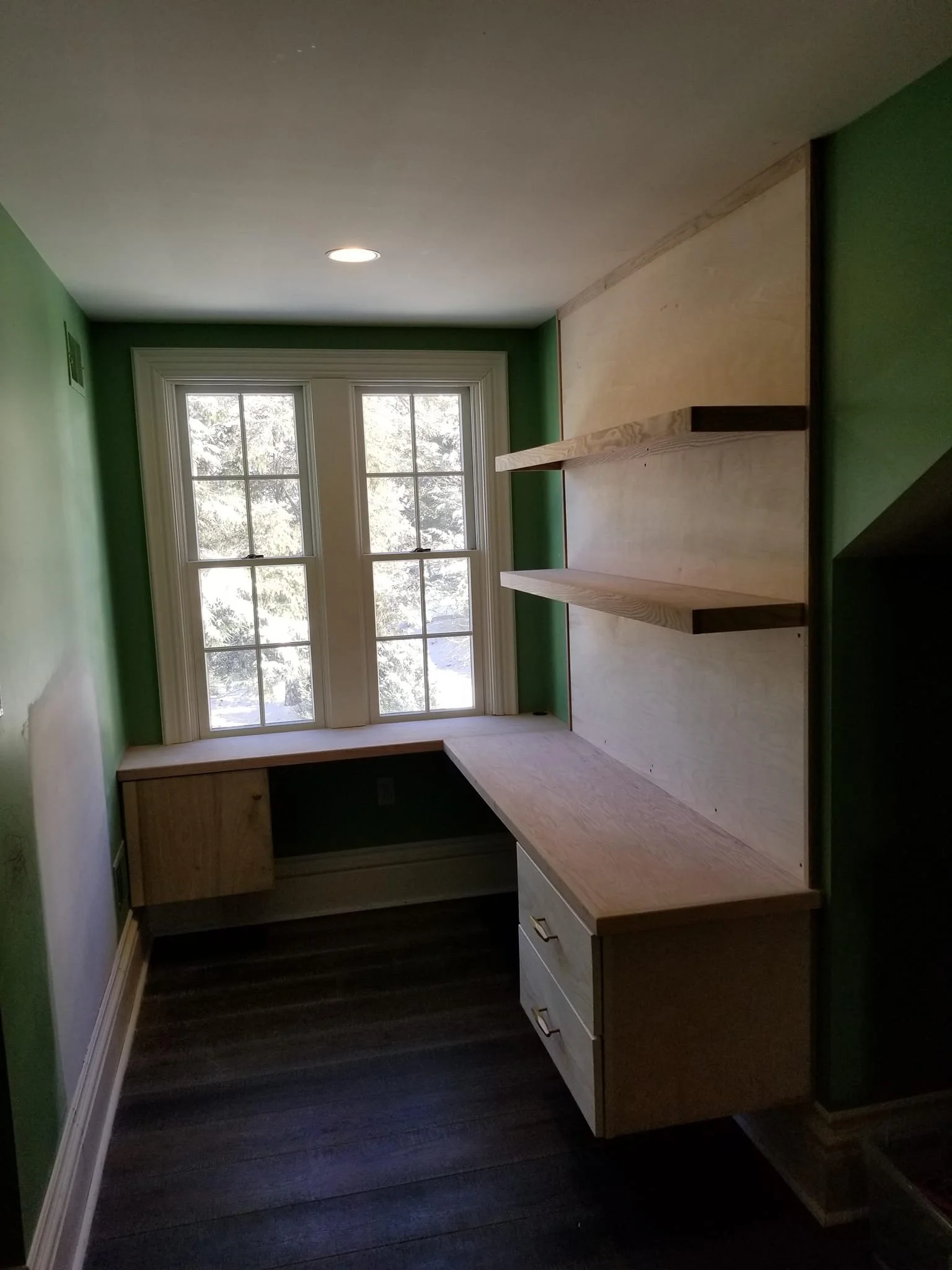 Room with green walls, large window with two sections, built-in wooden desk with drawers, and wooden shelves on the wall.