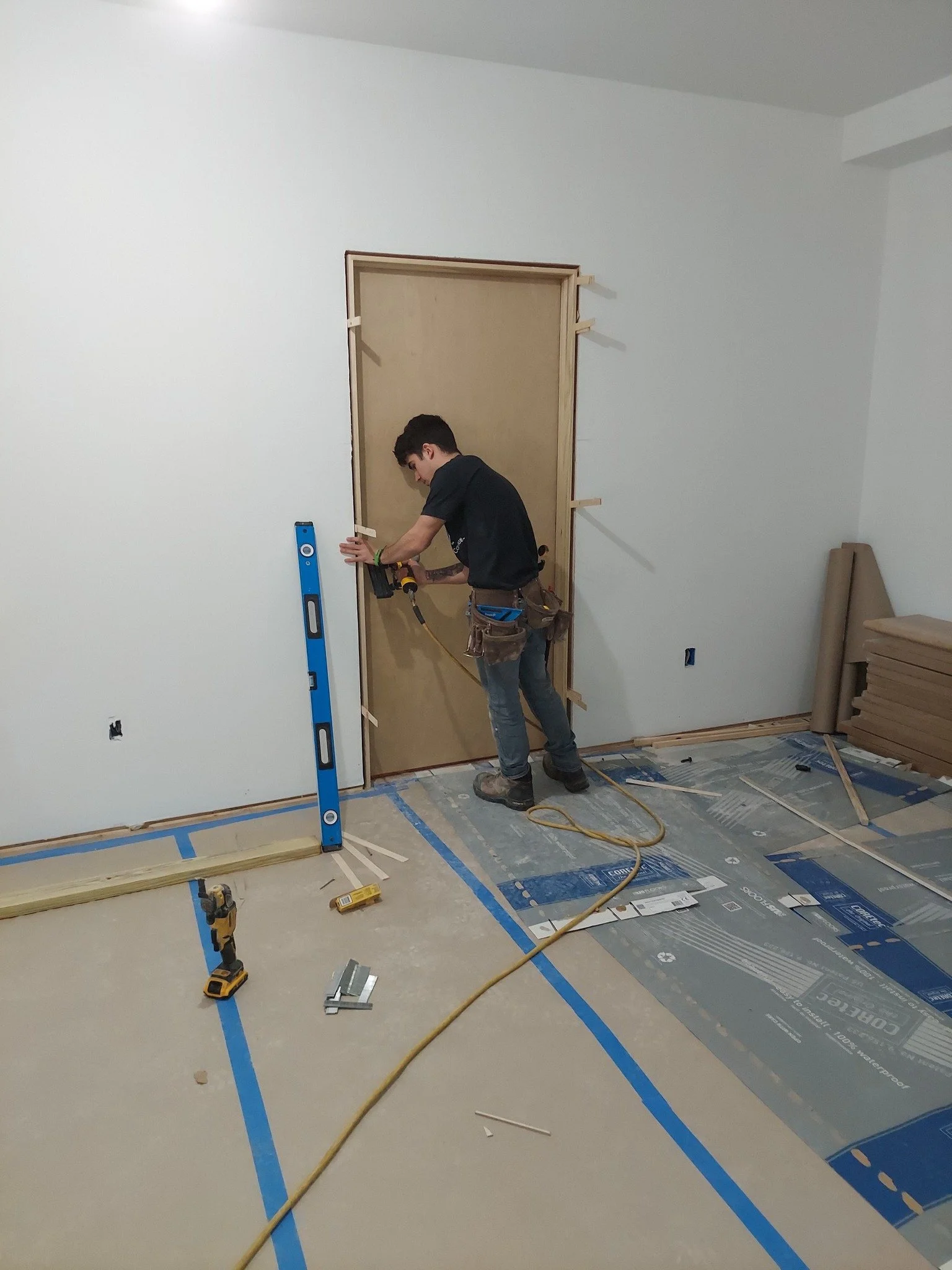 A man installing a door uses a drill, standing on a floor covered with protective material and construction tools, with a blue level and scattered hardware around him.