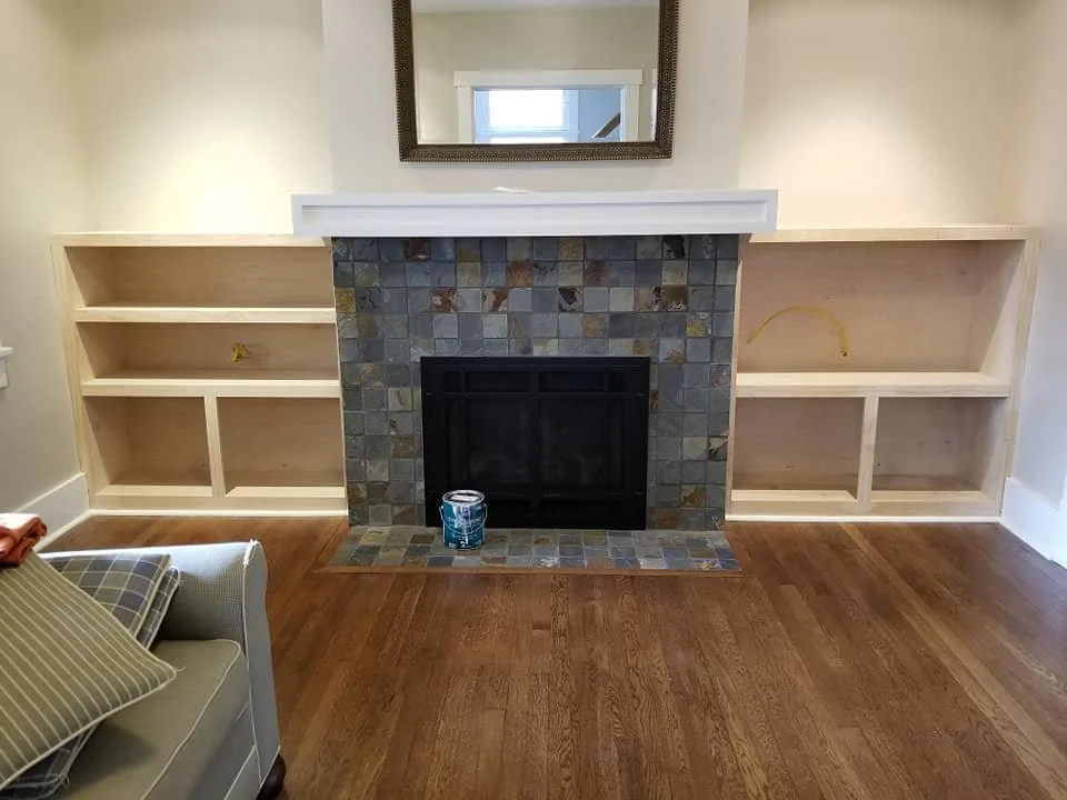 Living room fireplace with tile surround and built-in white shelves on either side. Mirror above the fireplace, and a can of paint on the floor in front of it.
