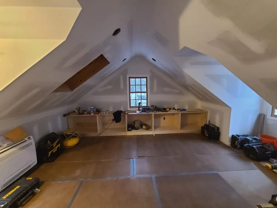 Attic space with sloped ceilings, drywall in progress, and a small window in the back wall, with construction tools and equipment scattered around.