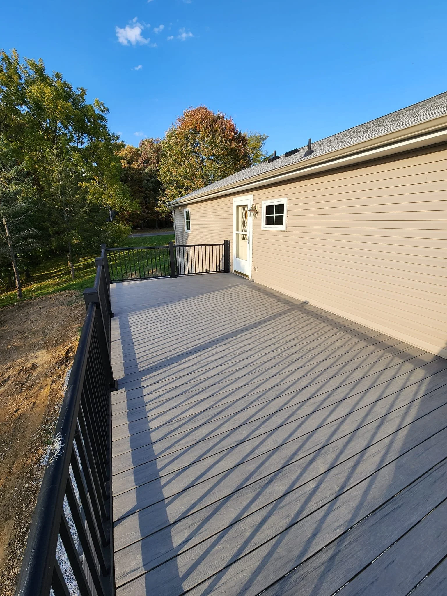 A spacious outdoor balcony with gray wooden flooring and black metal railing, attached to a beige house with a white door and small window, surrounded by trees with autumn foliage under a clear blue sky.