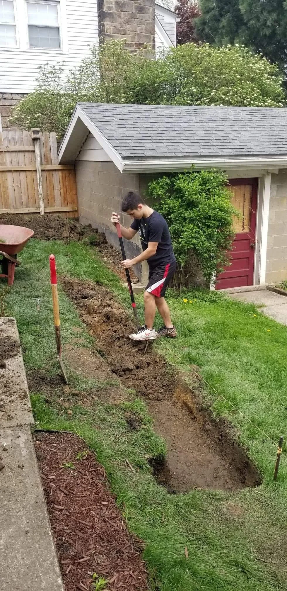 A young man is digging a trench in a backyard with a shovel and a pickaxe. There is a brick building and a small shed, with trees and grass around.