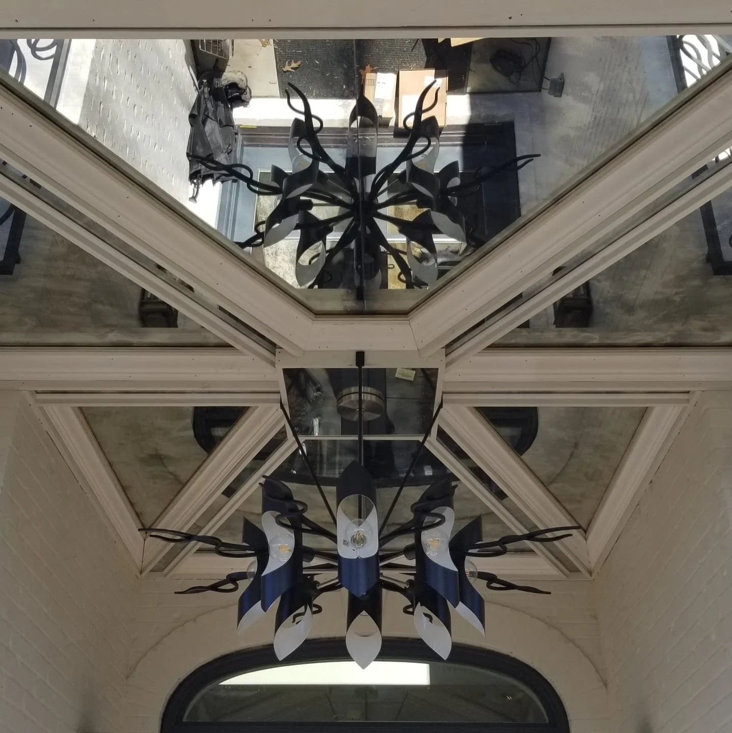 View of a chandelier hanging from a ceiling with mirrored panels, reflecting the fixture and part of the room below.