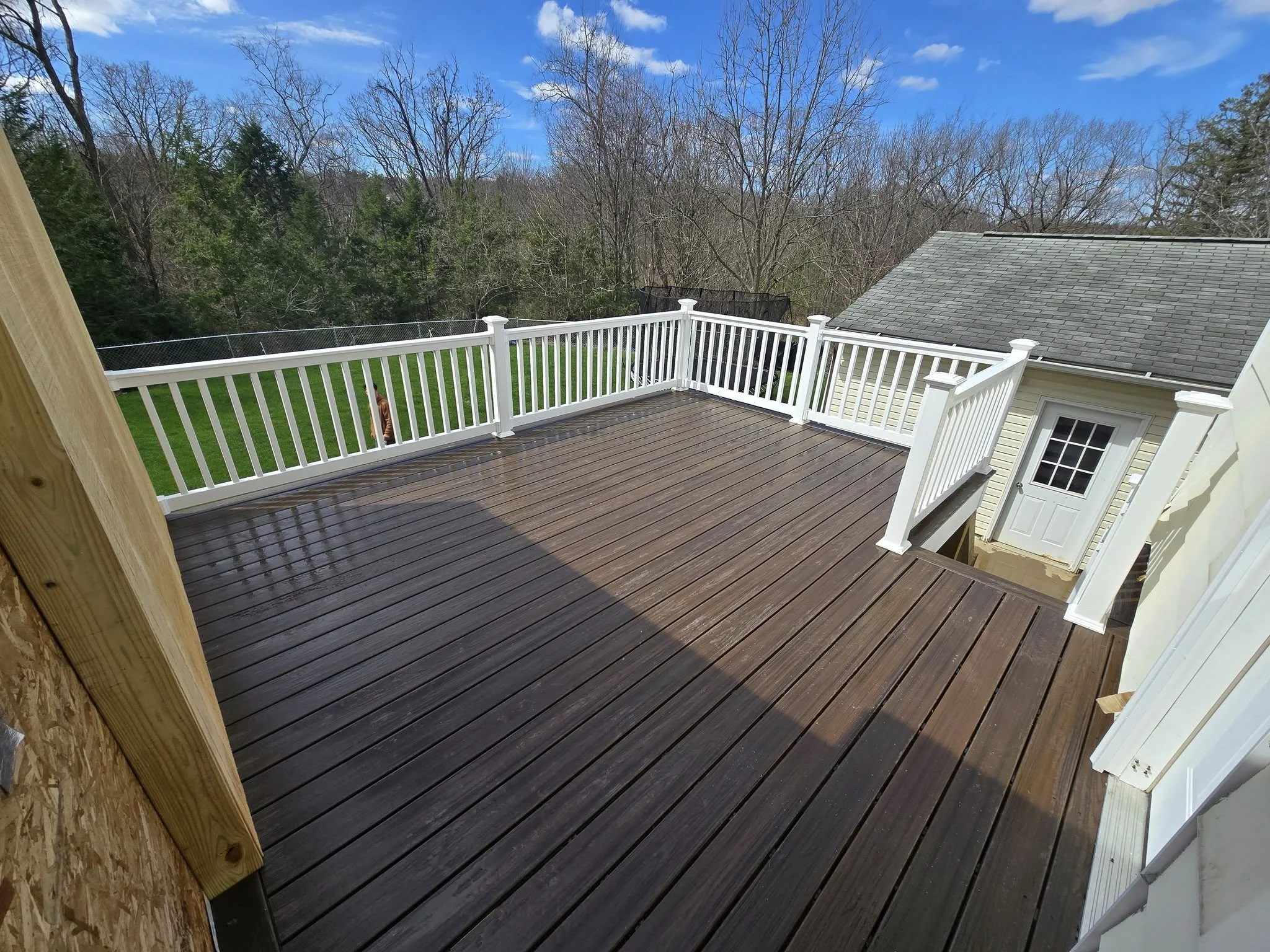 View of a wooden deck with white railing, attached to a house, overlooking a backyard with green grass and trees, under a partly cloudy sky.