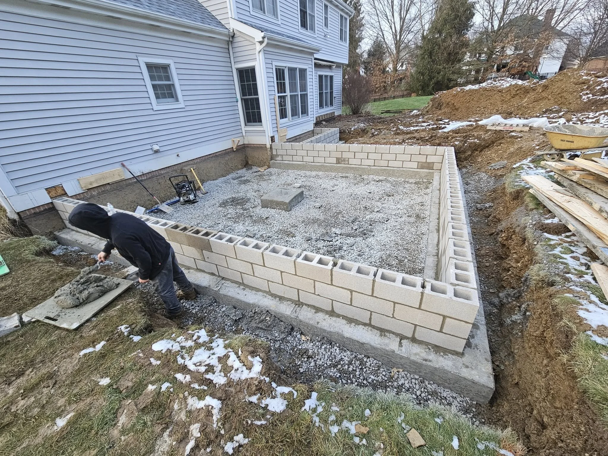 A construction worker building a cinder block wall around a backyard patio or garden area. The area is partially frozen with some snow visible on the ground and nearby dirt mounds.