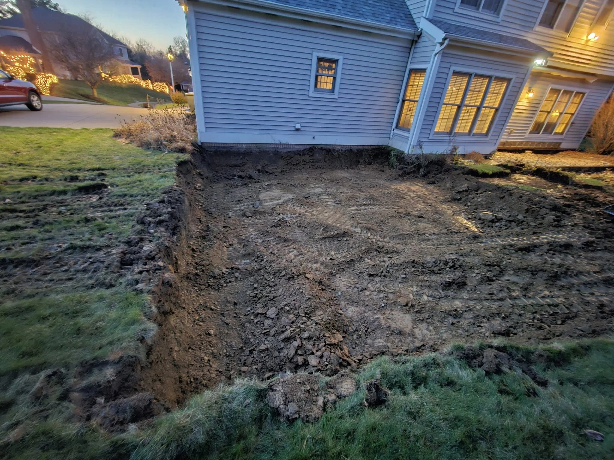 A backyard with a lawn on the left and a dug-out area near the house on the right, with a house with lit windows in the background.