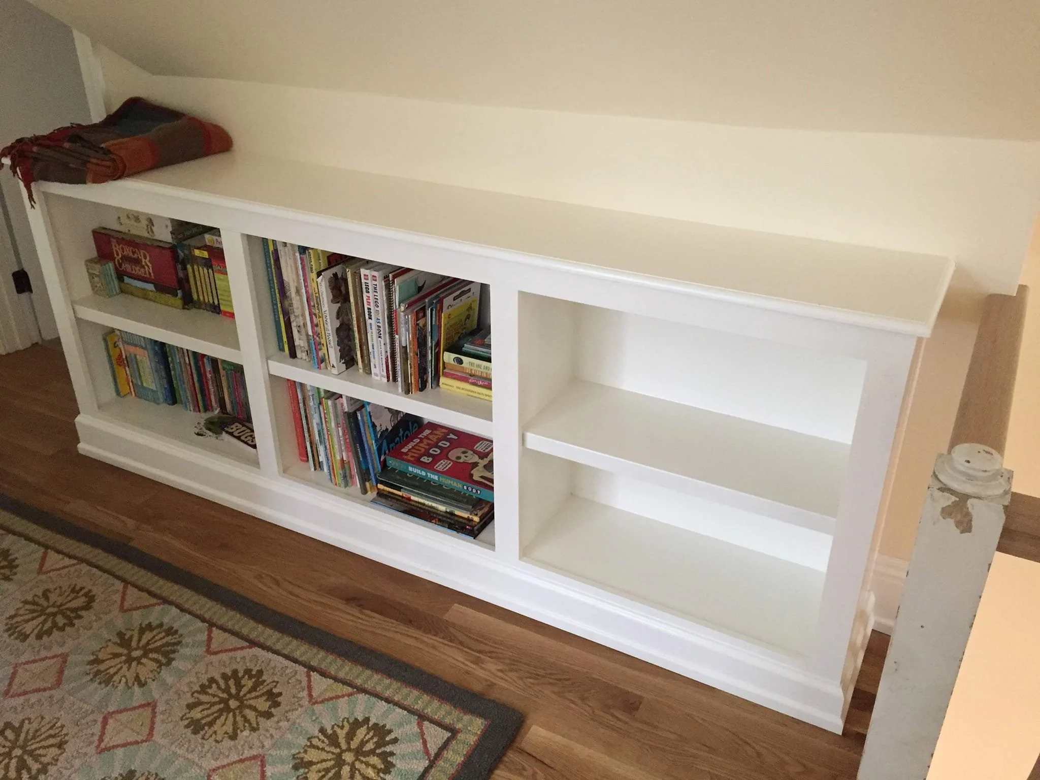 A white bookshelf with three shelves filled with books on the left side and two empty shelves on the right side, placed on a wooden floor with a patterned rug partially visible in front.
