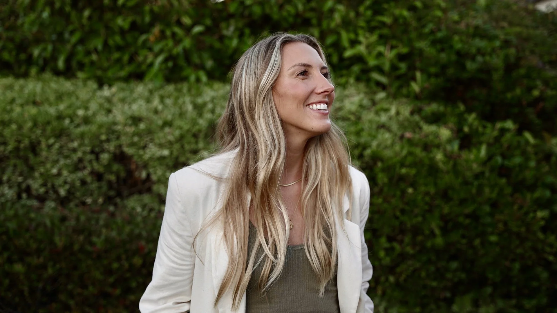 A woman with long blonde hair and a white blazer smiling outdoors in front of green shrubbery.