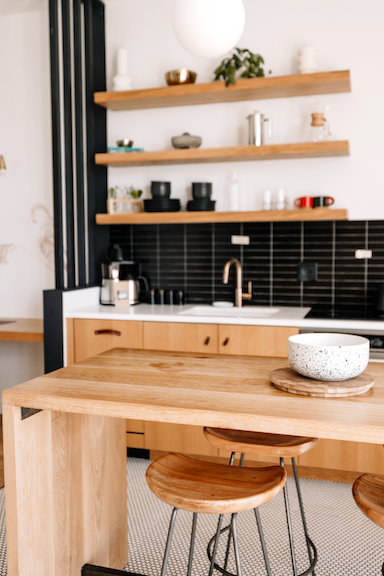 Organized kitchen with wooden island, black tile backsplash, open wooden shelves with decor, and bar stools