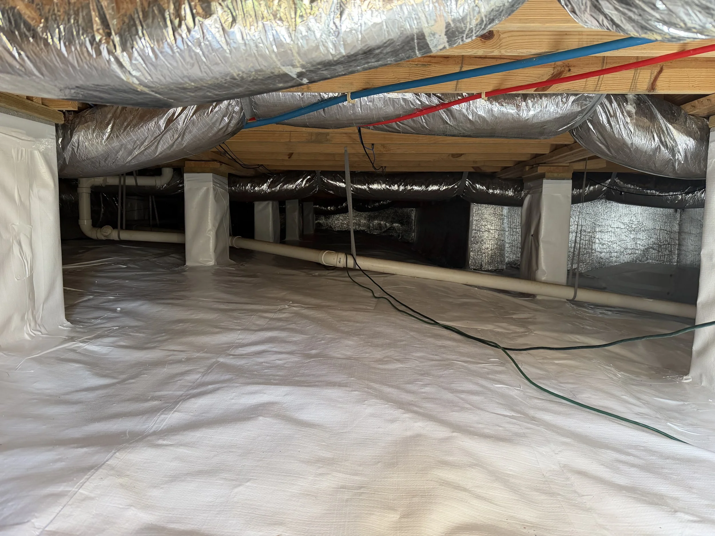 Indoor crawl space with ductwork, plumbing pipes, and wooden beams, covered with silver insulation foil and white vapor barrier on the floor.