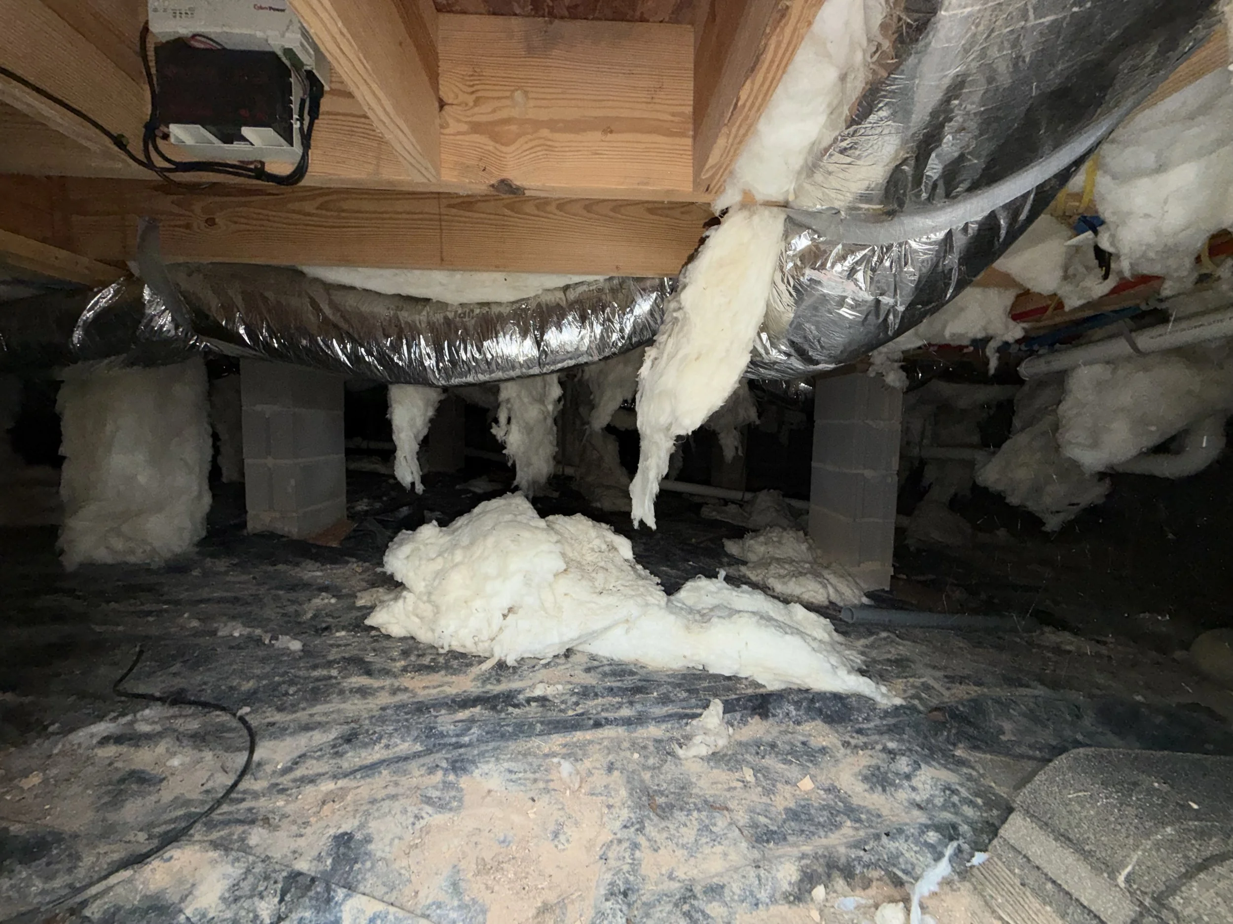 Crawlspace with ductwork, insulation hanging, damaged vapor barrier, and hanging electrical wiring.