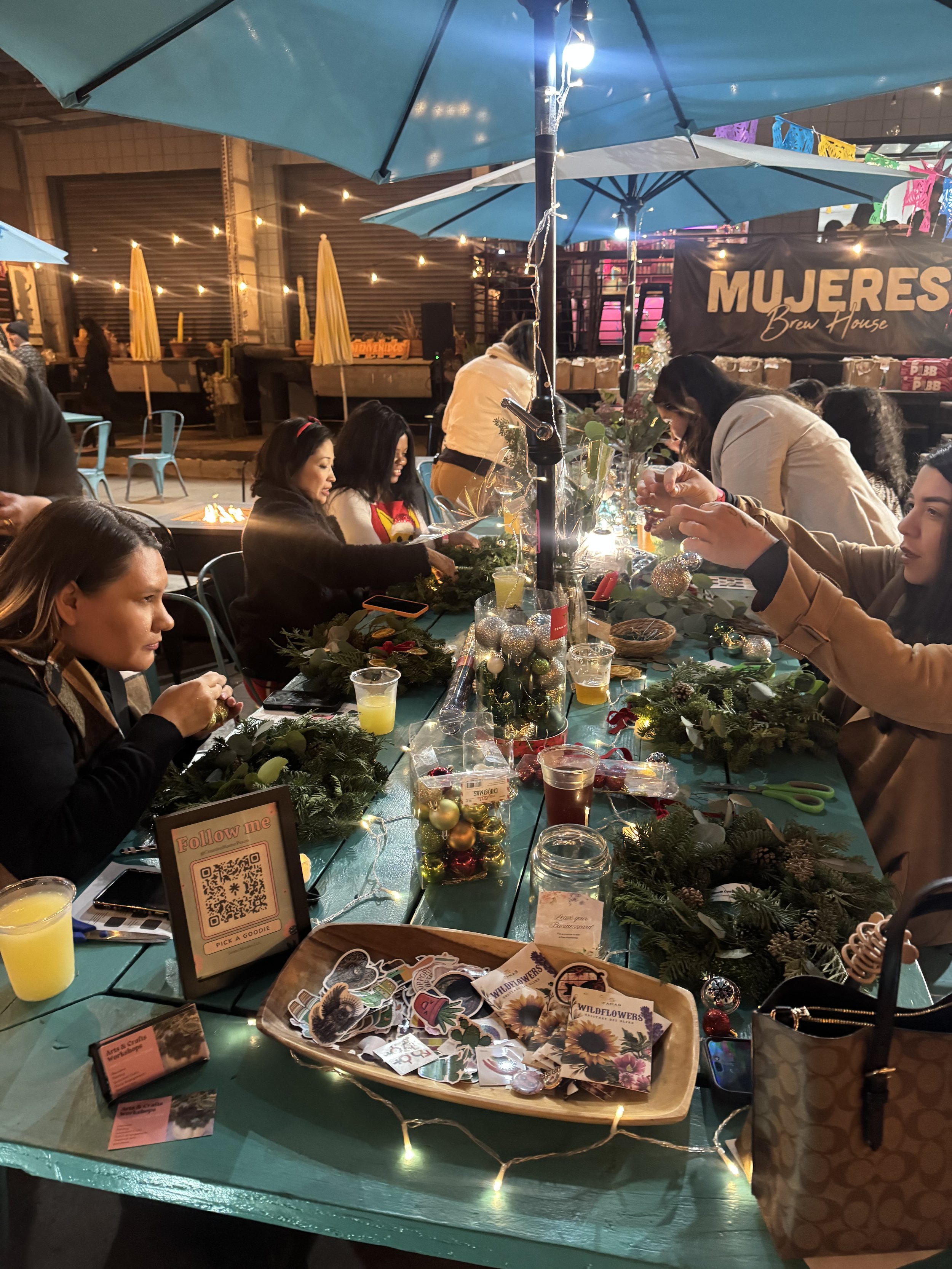 People crafting holiday wreath outside in the evening in a outdoor venue with string lighting and umbrellas