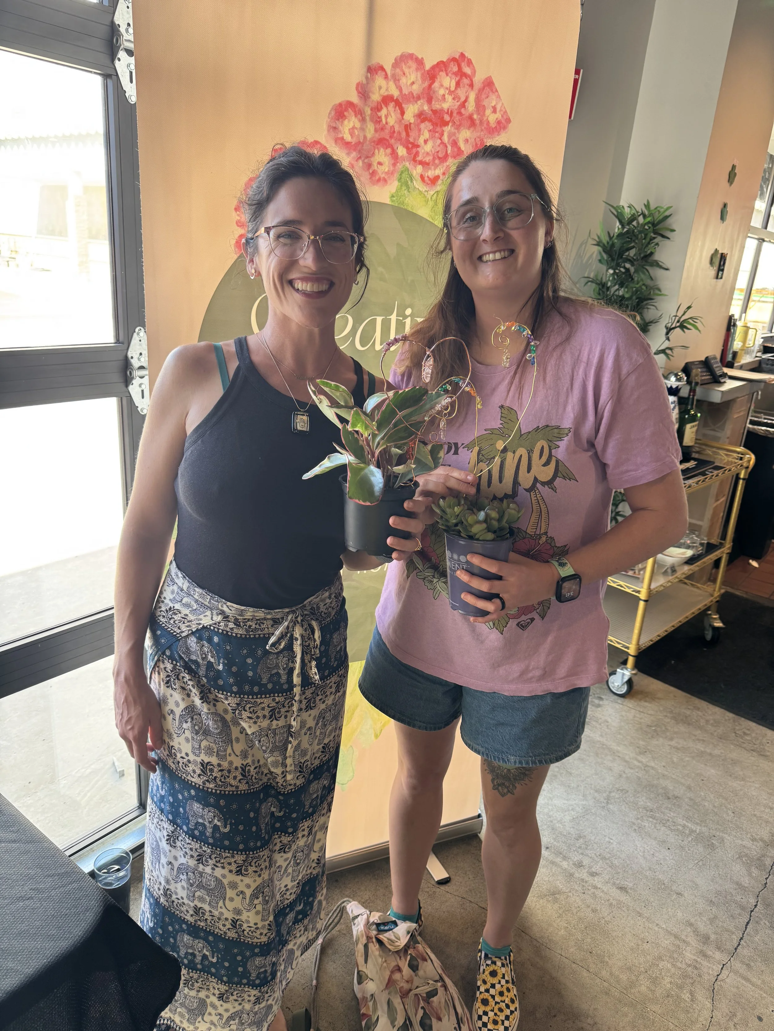smiling women holding plants with crafted beaded plant stakes in front of a creative master peaces banner