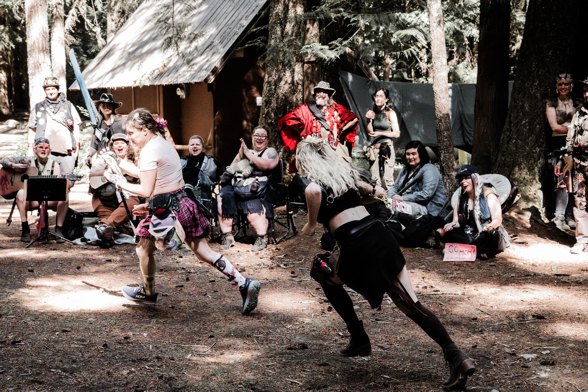 Group of people gathered in a wooded area, some sitting on chairs, while one person is running with a plastic sword and a helmet, and another person in black attire is running after them. The group appears to be participating in or watching a cosplay or themed event.