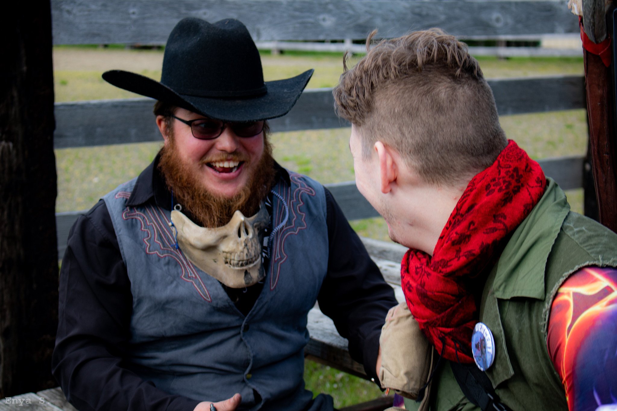 Two men sitting outdoors, laughing and engaging in a friendly arm wrestling match. One man is wearing a black cowboy hat, glasses, a skull necklace, and a dark shirt. The other man has short, faded hair, a red scarf, green jacket, and a button on his sleeve. They appear to be enjoying each other's company.