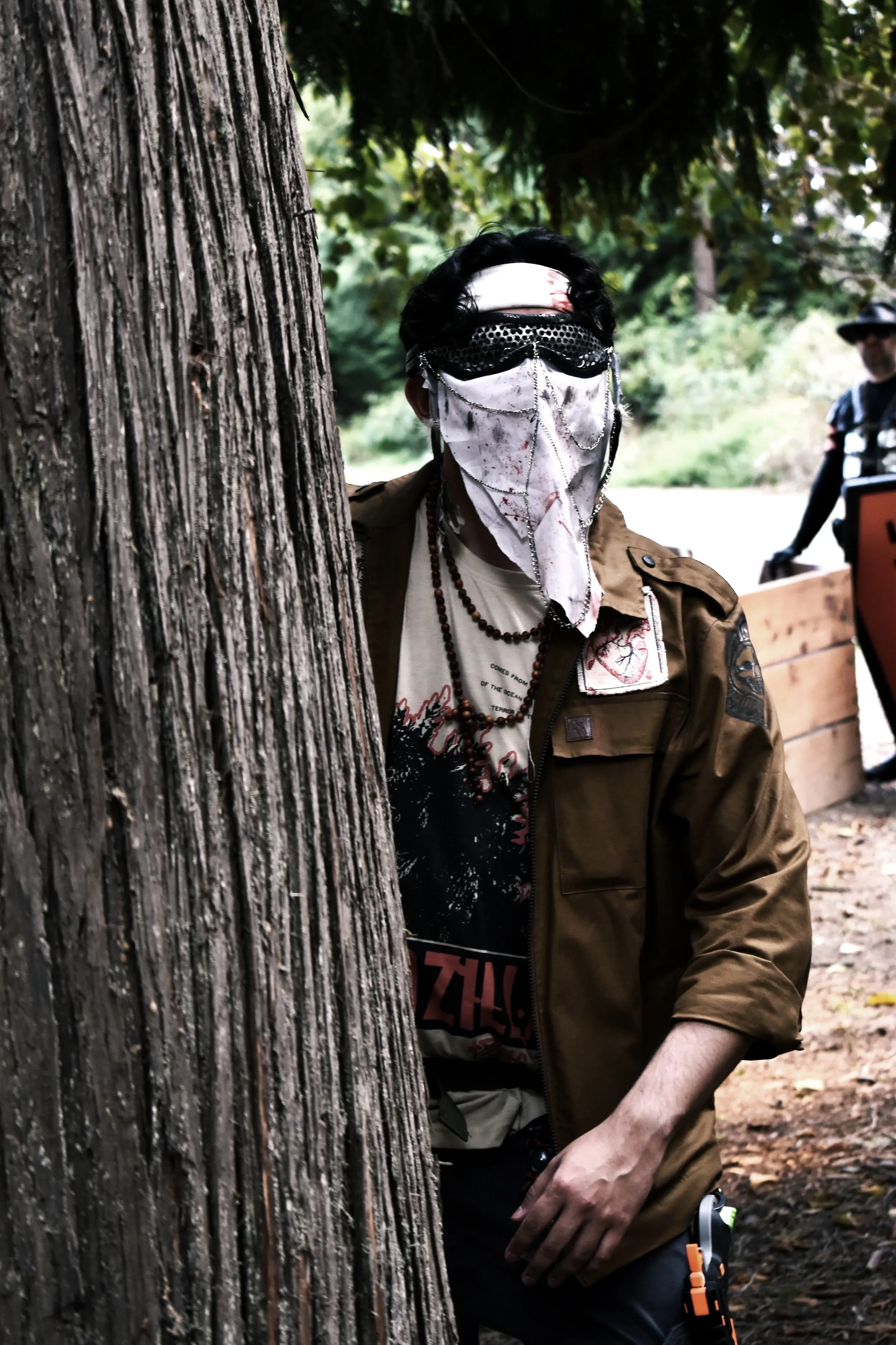 Person wearing a mask with chains, sunglasses, and a brown jacket hiding behind a tree in a wooded outdoor area.