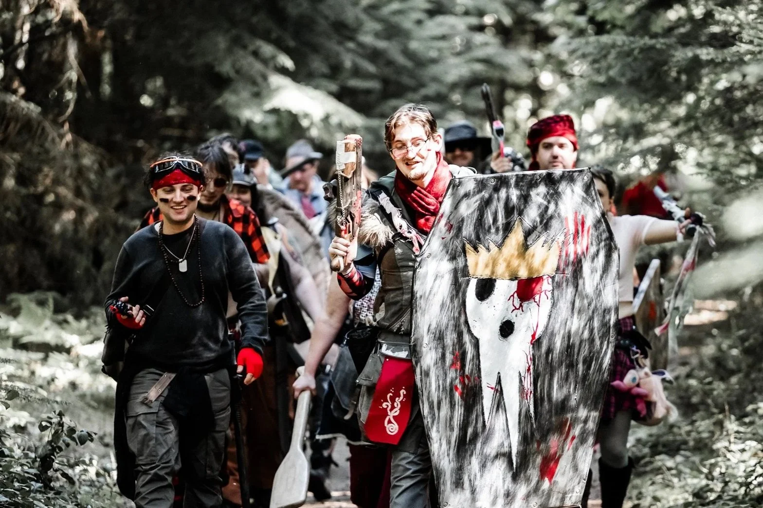 A group of people dressed in costumes, walking through a forest, some holding weapons and a painted shield with a skull design.