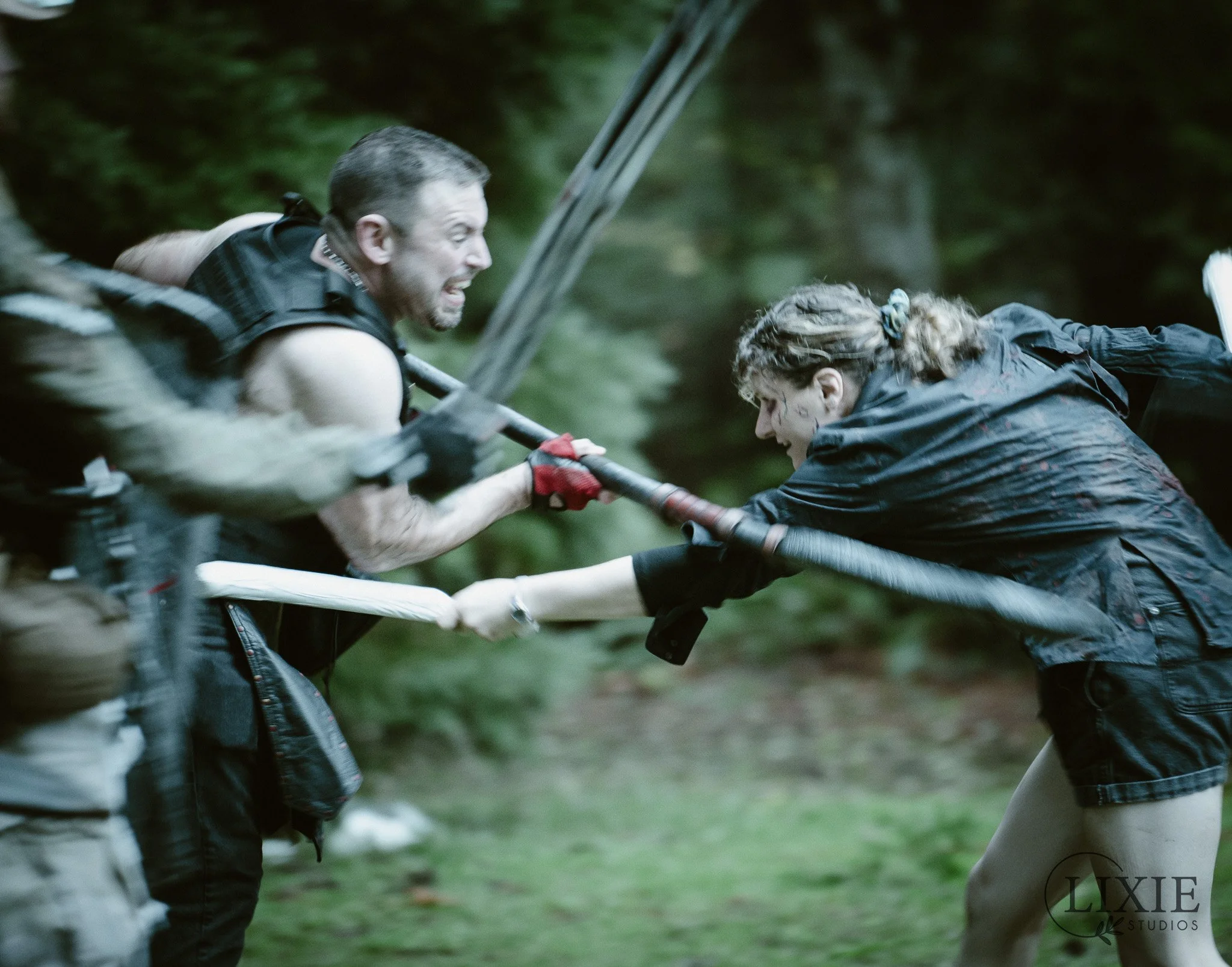 Two people engaged in a physical struggle or fight in a forest, grabbing each other's arms or holding a long stick, with intense expressions.