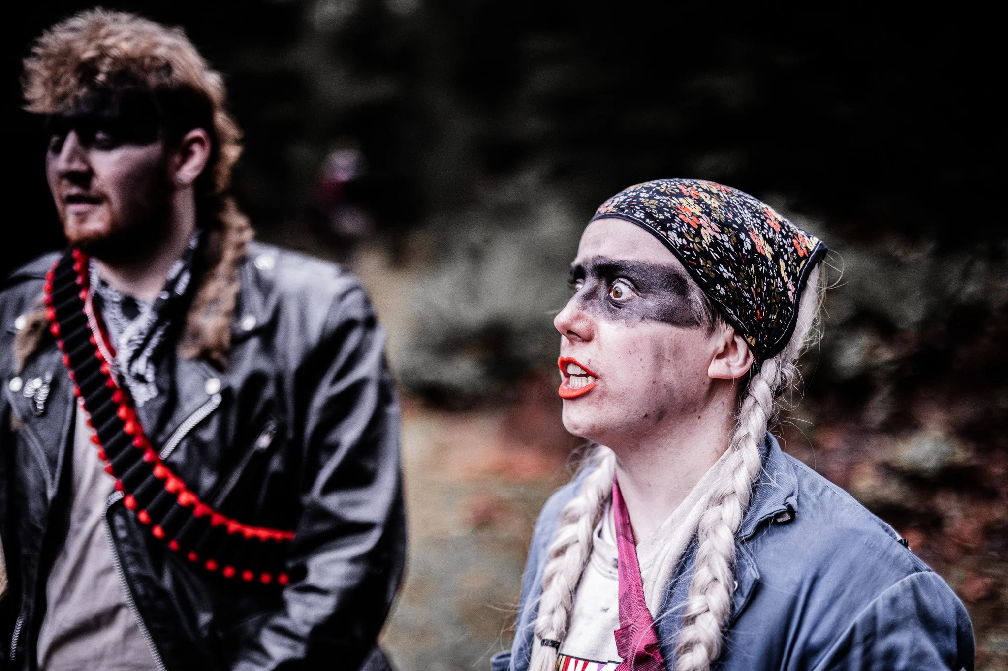 Two people dressed in costumes with makeup, one with dark face paint and braided hair, appears to be angry or shouting, while the other wears a leather jacket with a bandolier of red pellets, against a blurred background outdoors.