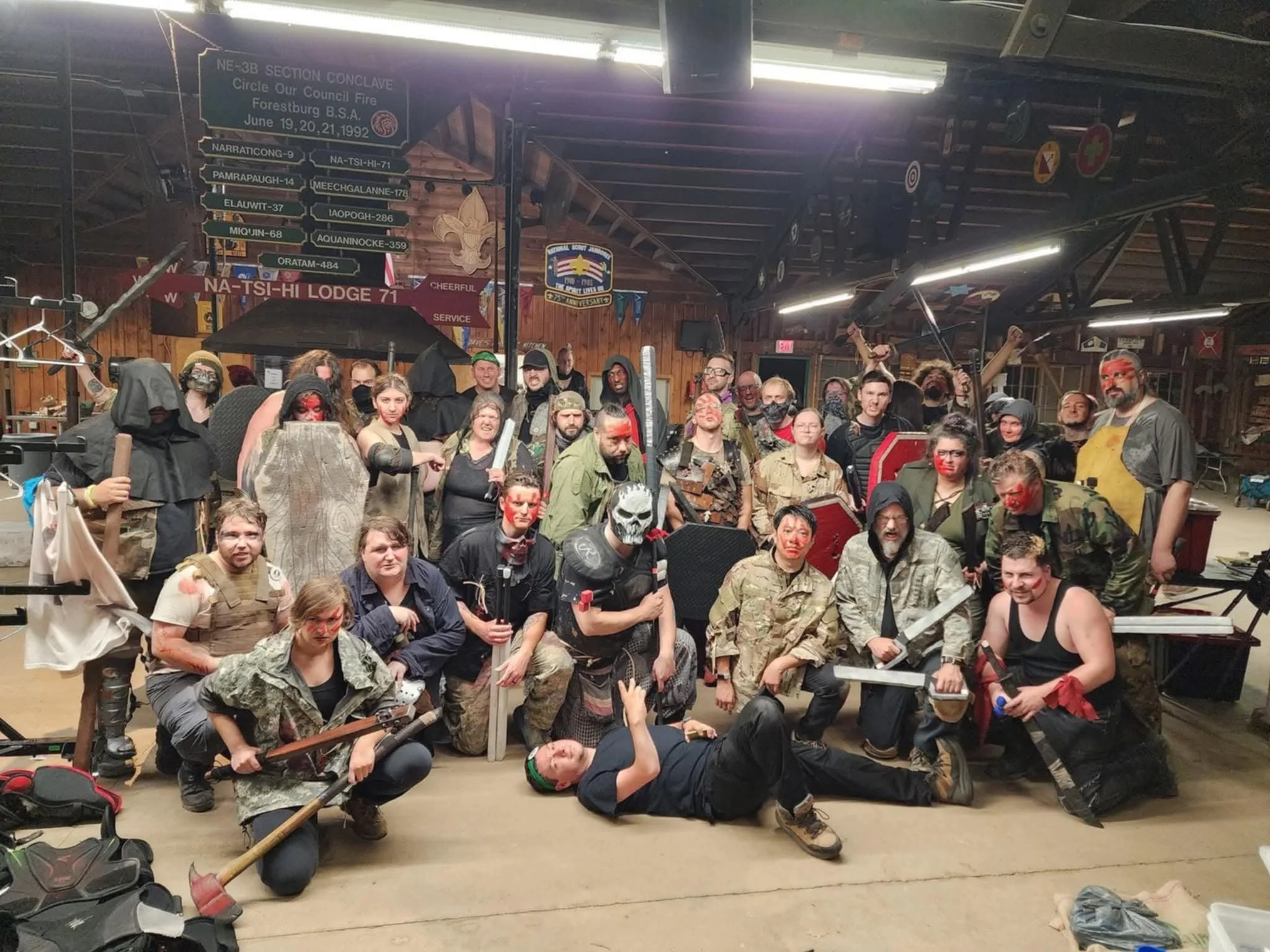 A large group of people dressed in cosplay costumes, many with face paint and weapons, gathered indoors with wooden walls and ceiling, some wearing military or fantasy-themed outfits.