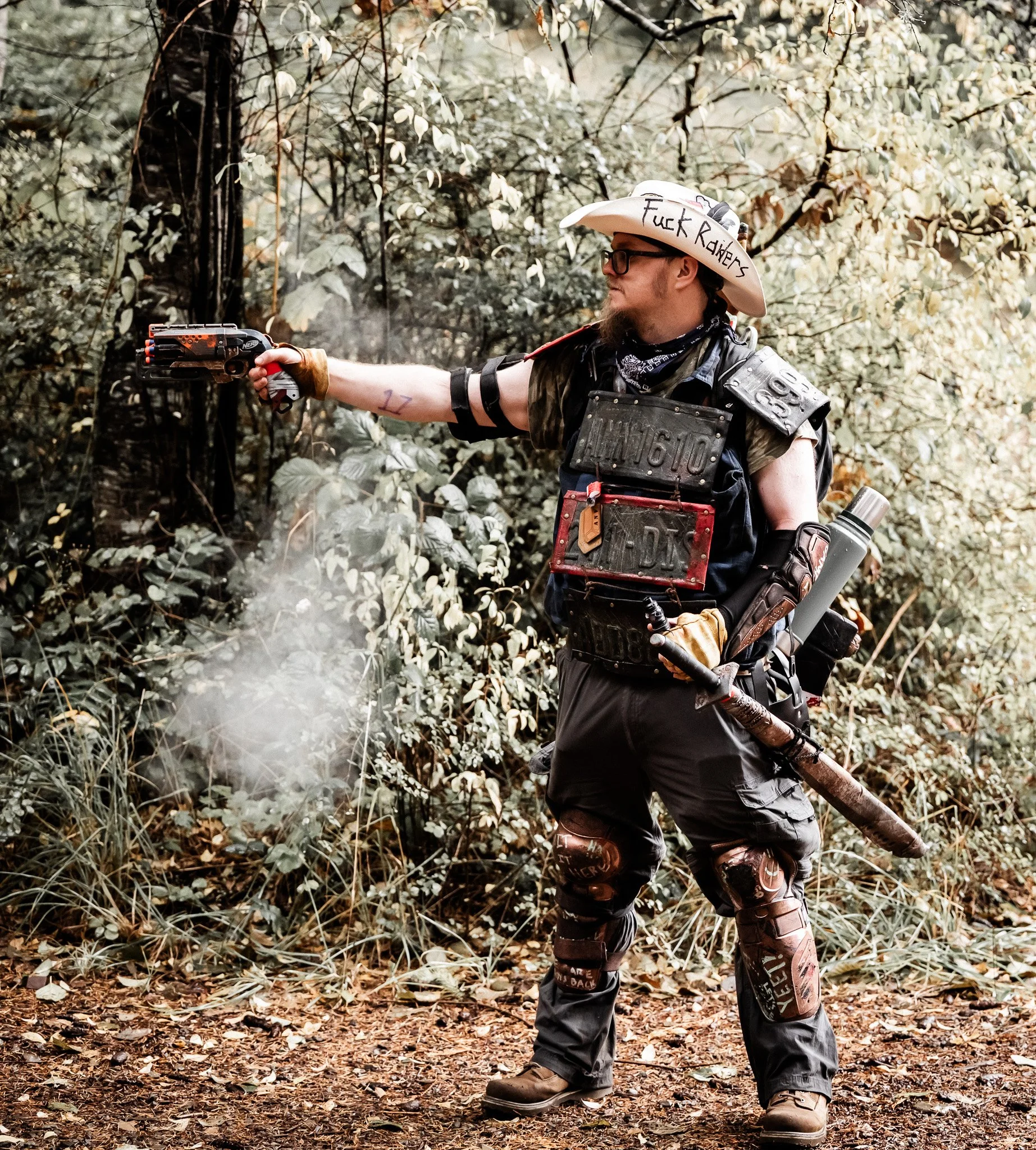 A person dressed in post-apocalyptic or cosplay attire, wearing a cowboy hat with the words 'Fuck Raiders' written on it, firing a gun in a forest setting.
