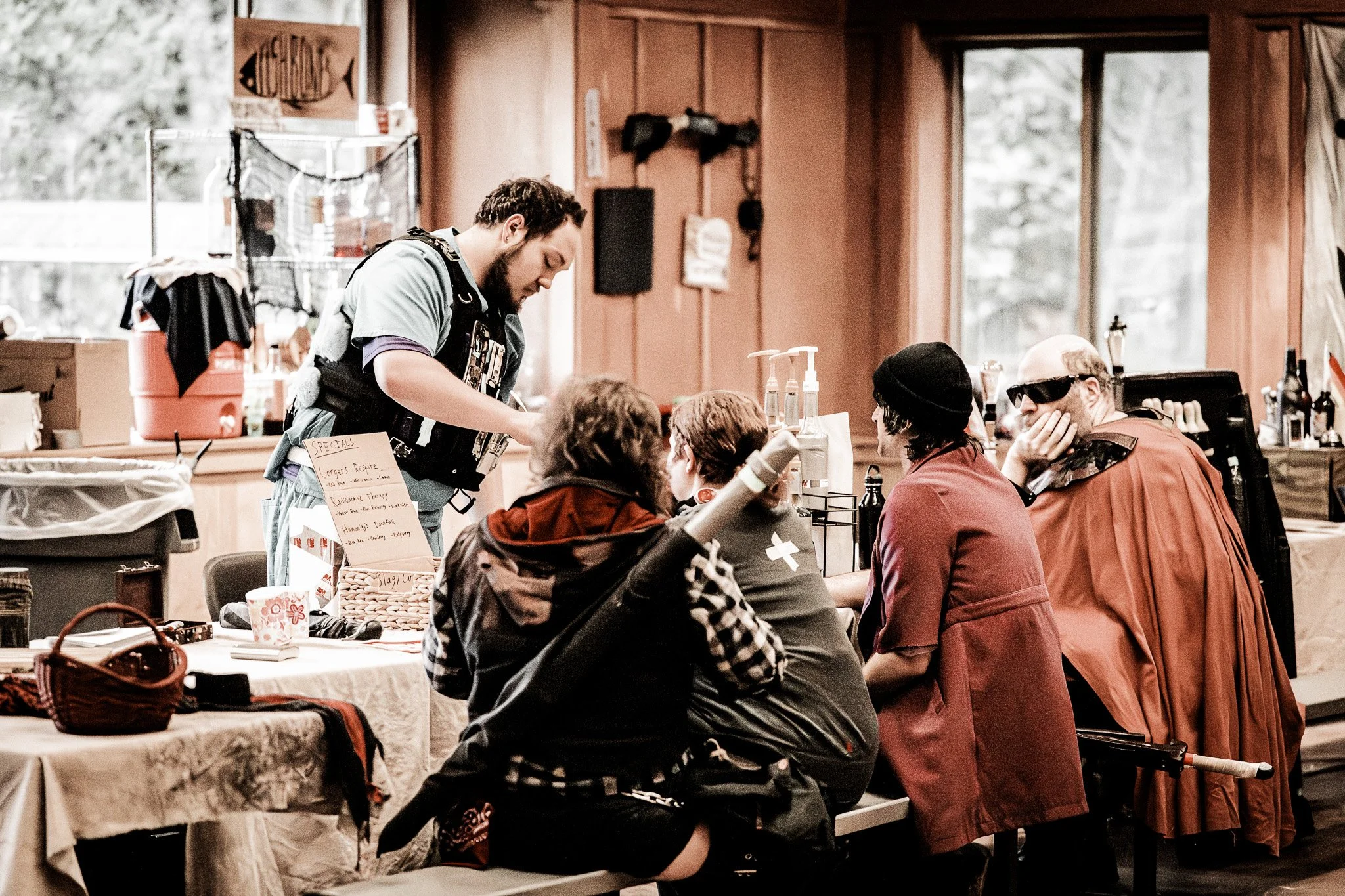 People sitting at a table in a casual indoor setting, with a man standing and engaging with them. The scene appears to be in a cozy, rustic space with wooden walls and large windows.