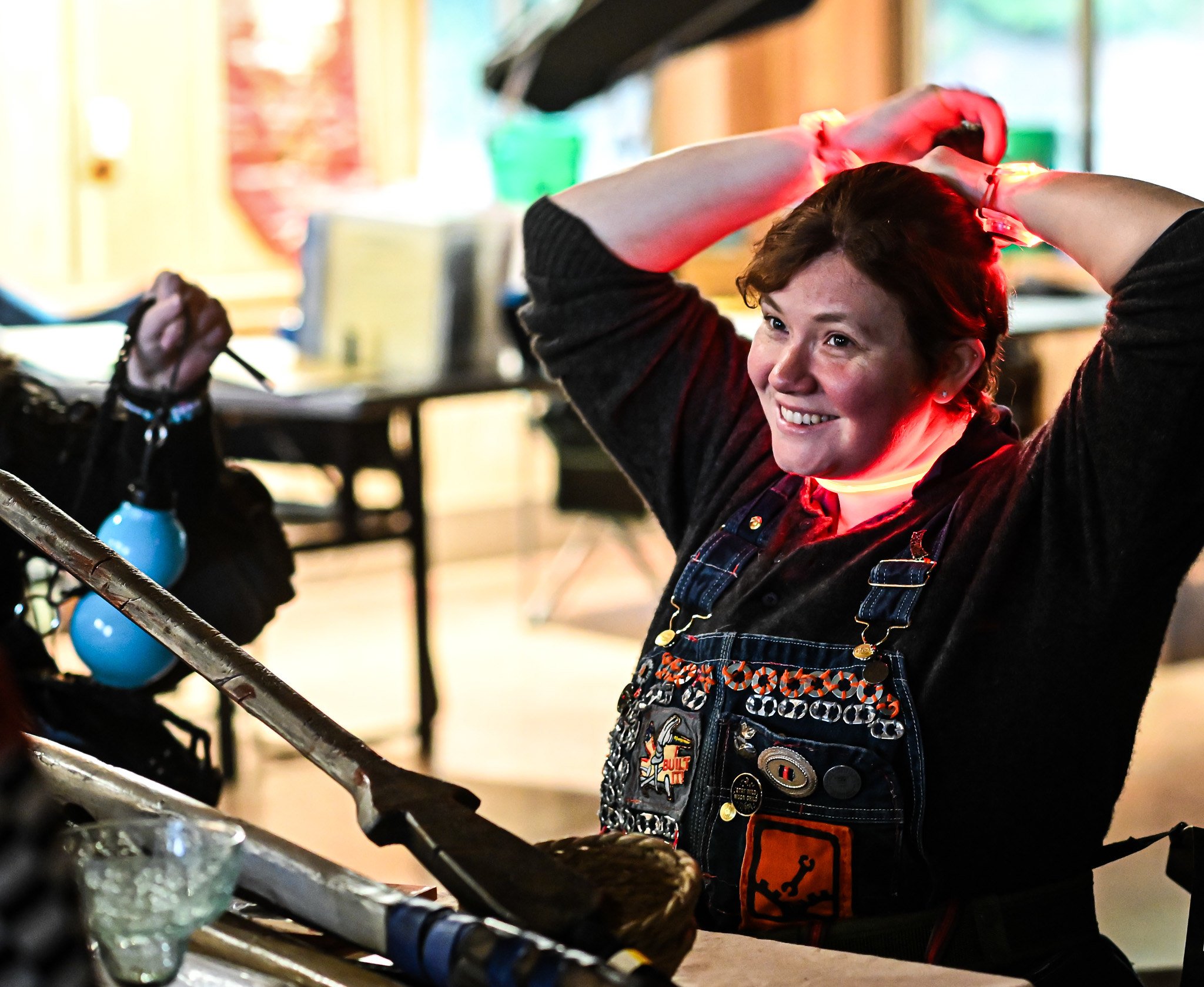 A woman with short brown hair, smiling, wearing a black outfit and a denim apron with patches, has her hands raised behind her head, standing in a room with various objects and food items on a counter.