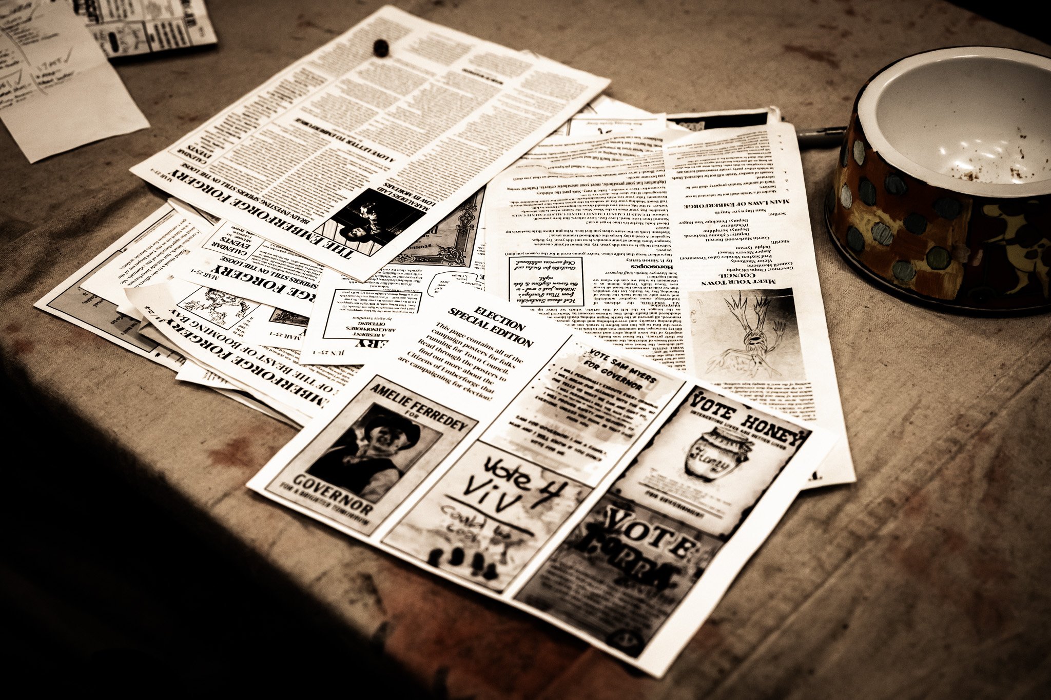 A table covered with scattered newspapers and flyers related to voting and political campaigns, and an empty pet food bowl on the right.