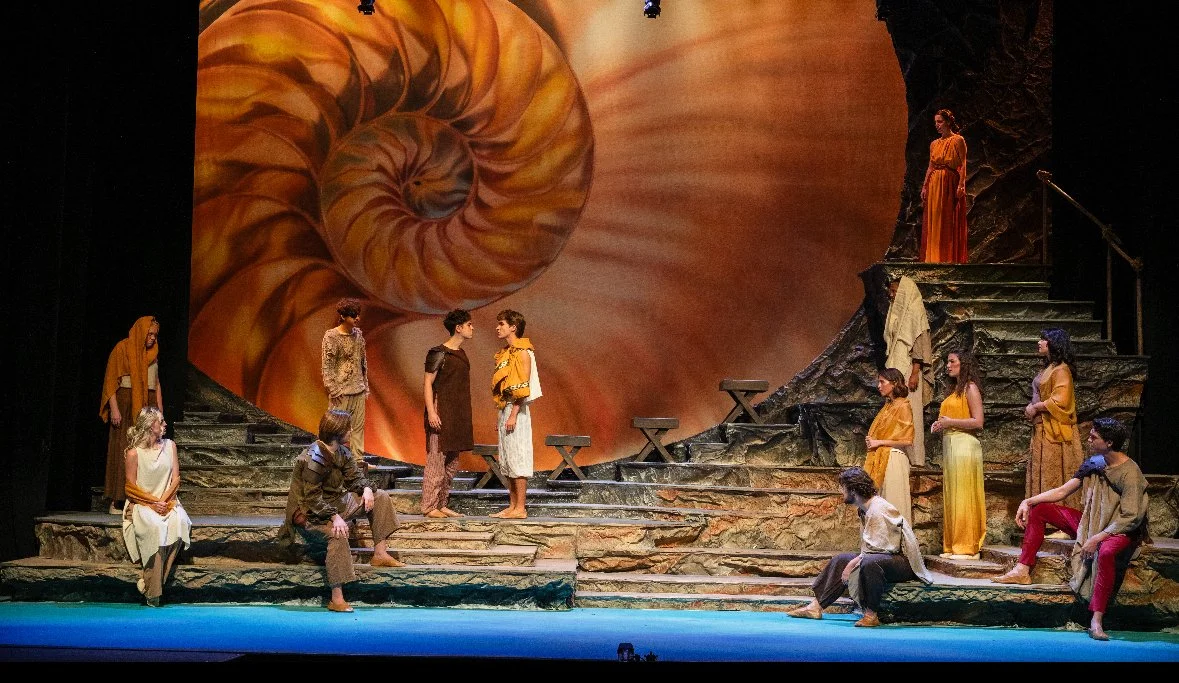 A theatrical stage scene resembling a nature-inspired setting with stone-like steps and a large spiral shell backdrop. Multiple actors dressed in earthy-toned costumes are performing, with some standing on stairs, others seated or crouched, engaging in a dramatic scene.