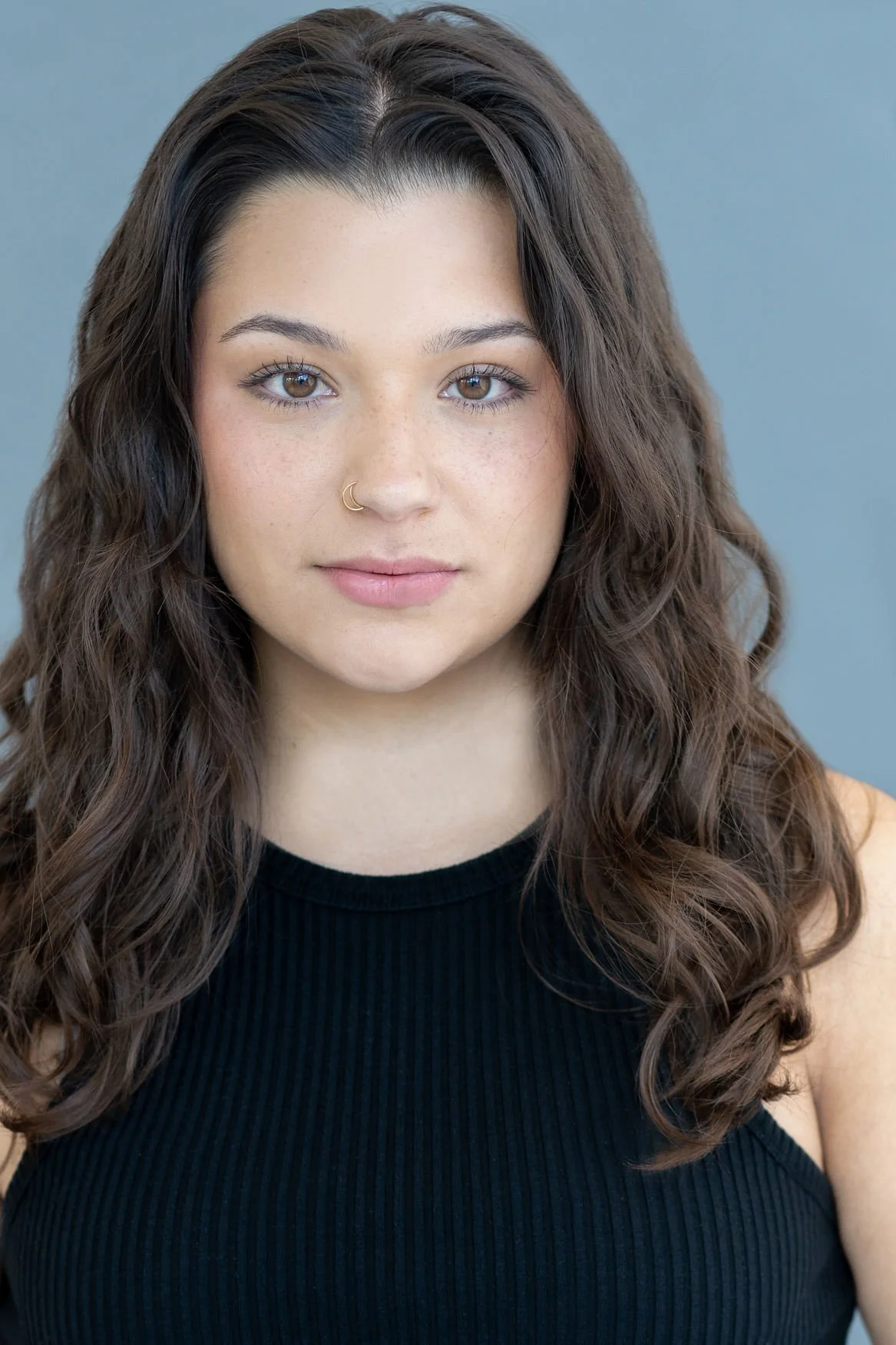 Close-up portrait of a young woman with brown wavy hair, hazel eyes, light makeup, and a nose ring, wearing a black sleeveless top against a solid blue background.