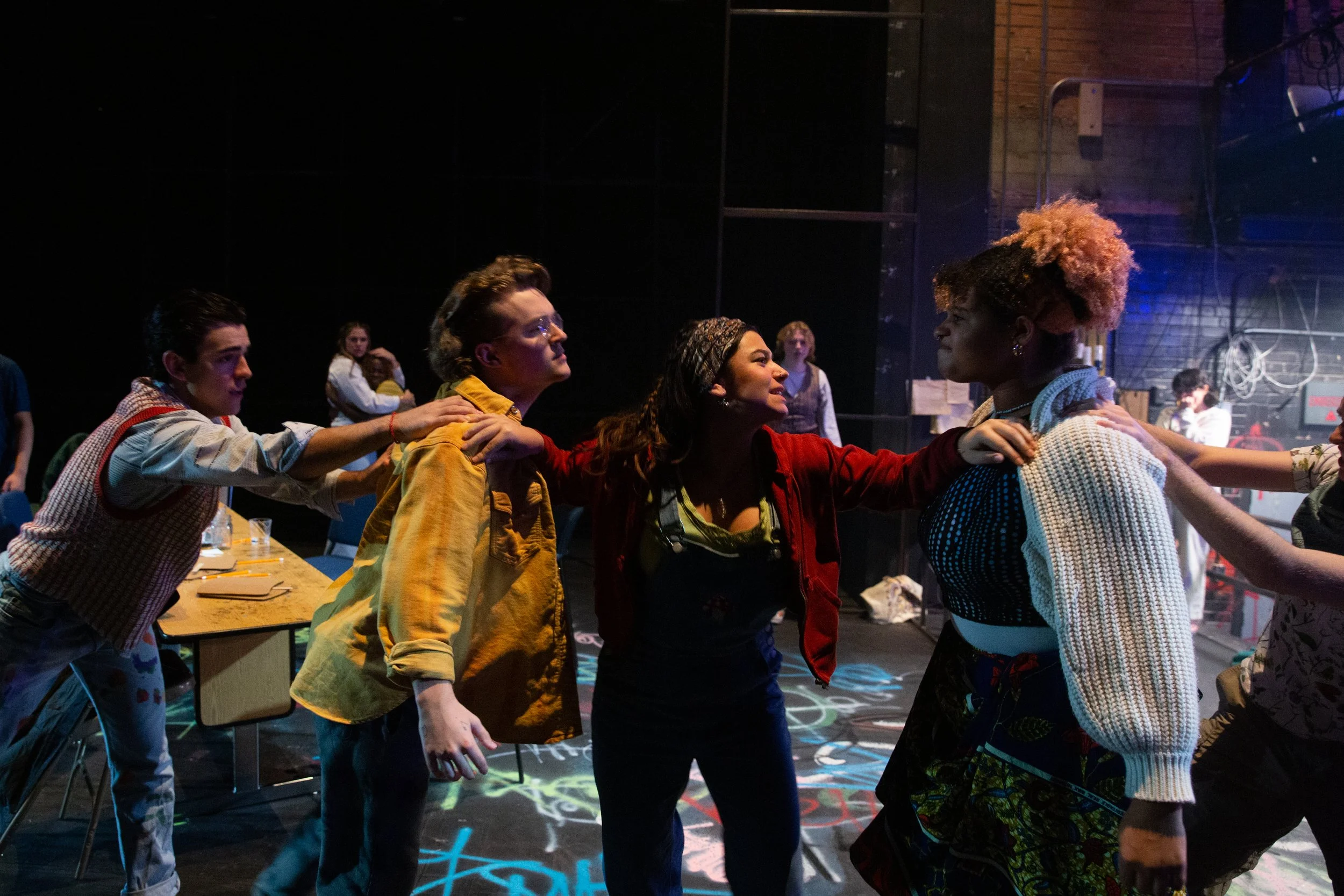 Group of people in a confrontation, with four women in the foreground and others in the background, in a dimly lit room with colorful lighting.