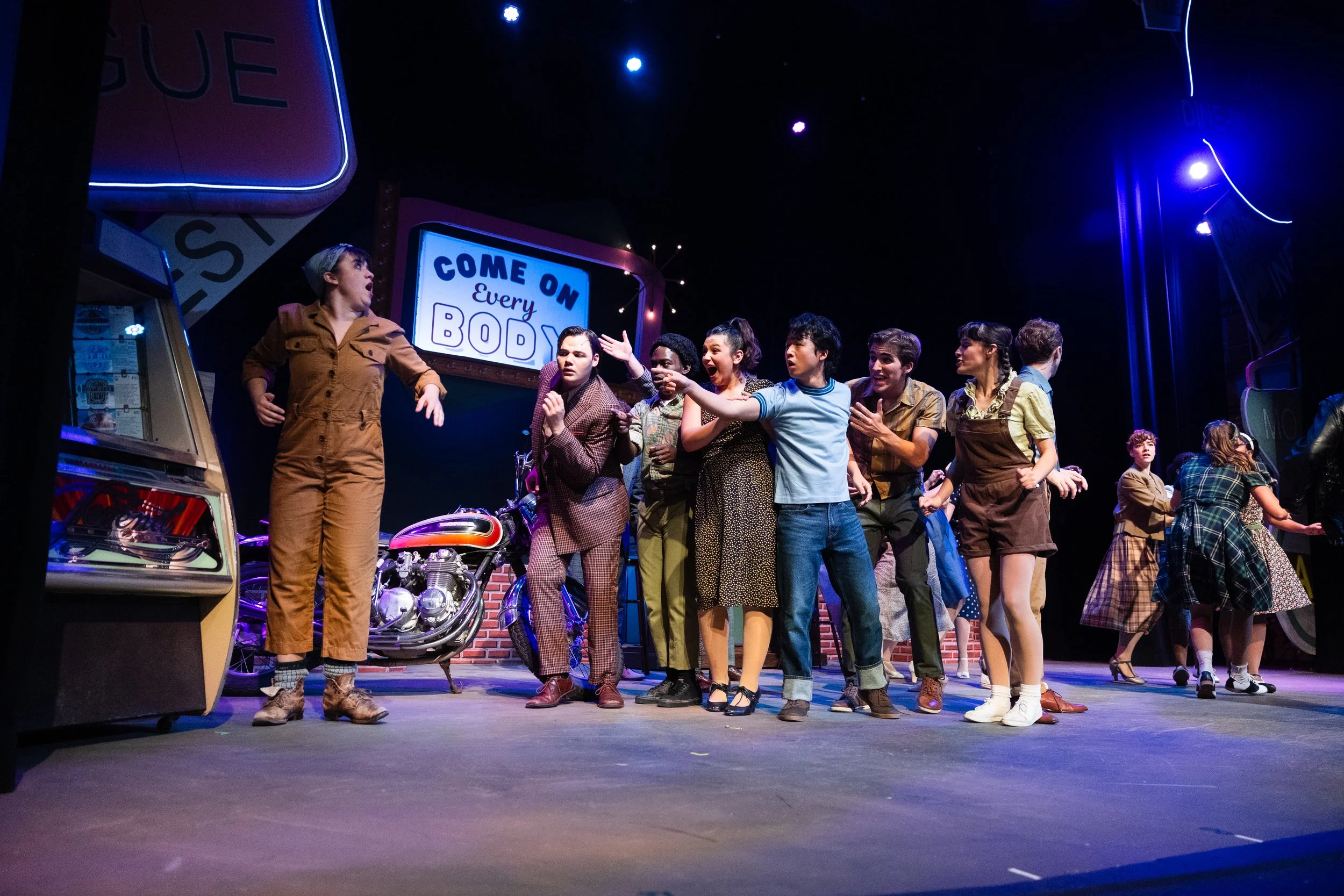 A group of young actors performing on stage in a theatrical production, with a neon sign in the background that says 'COME ON Every BODY' and a motorcycle prop.