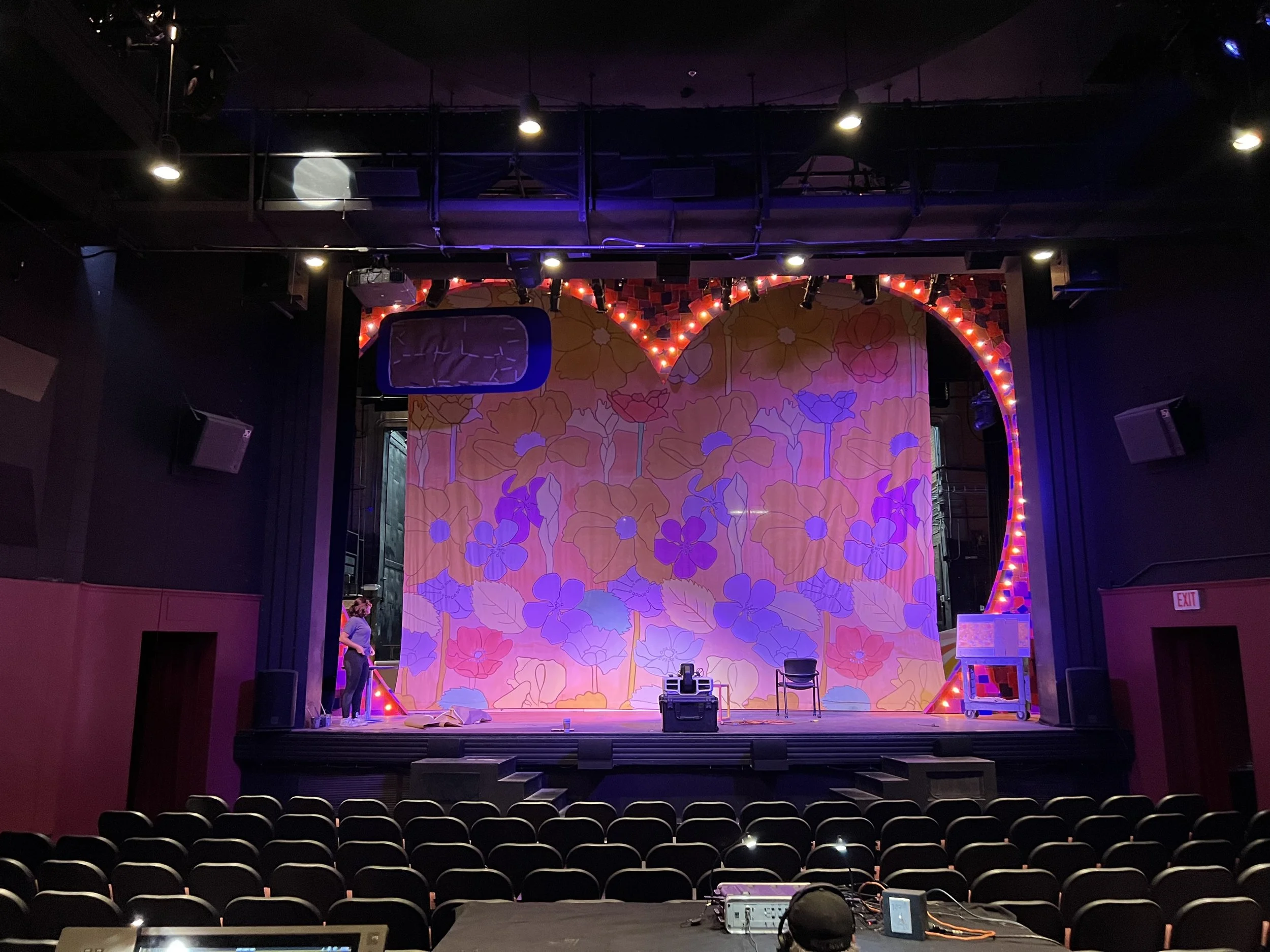 Empty theater stage with floral backdrop and decorative lights, with a person working on the left side and theatre seats in the foreground.