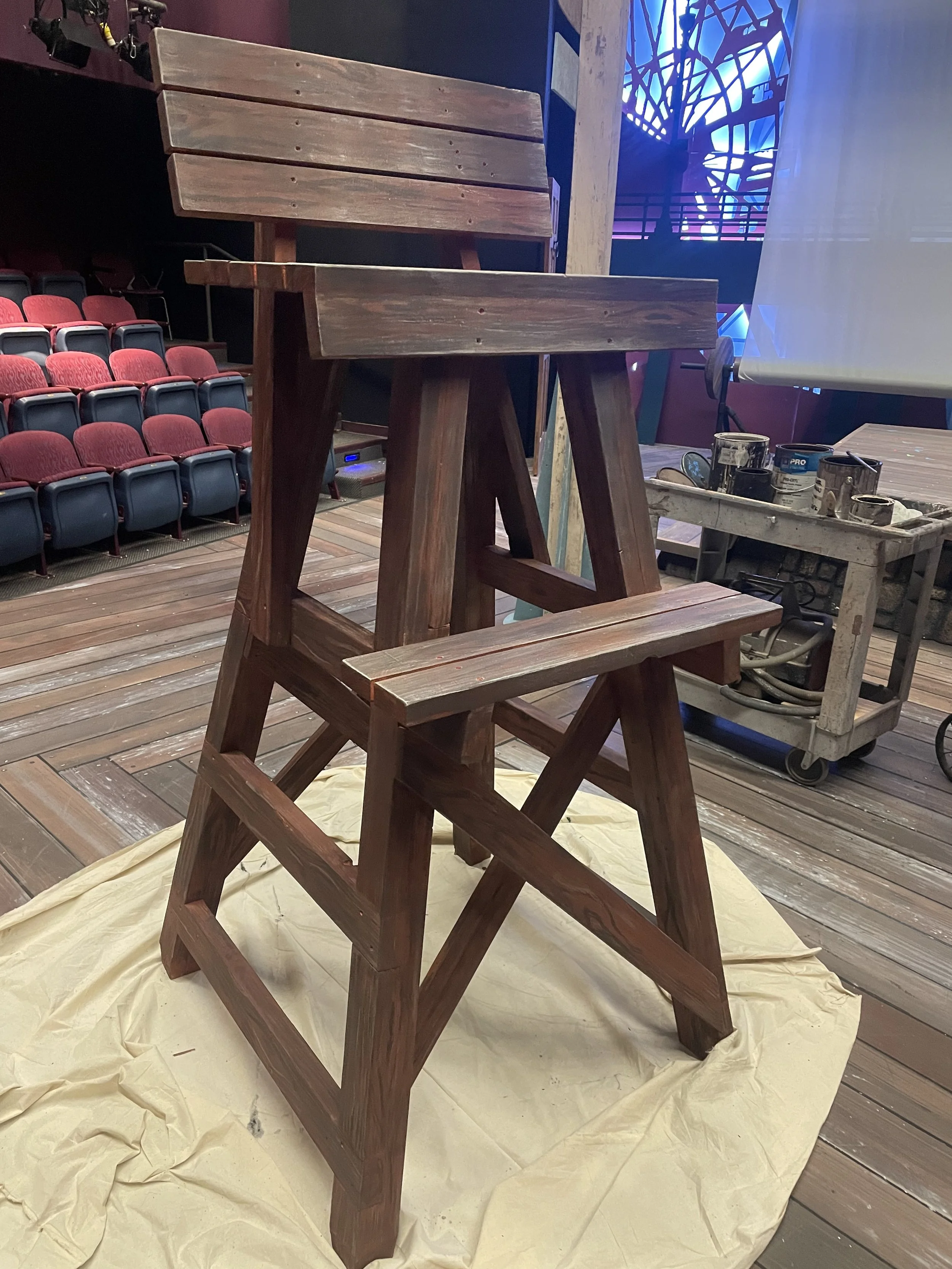 A wooden chair with a unique design featuring a bench-style seat and backrest, placed on a yellow cloth on a wooden floor in a studio or theater setting.