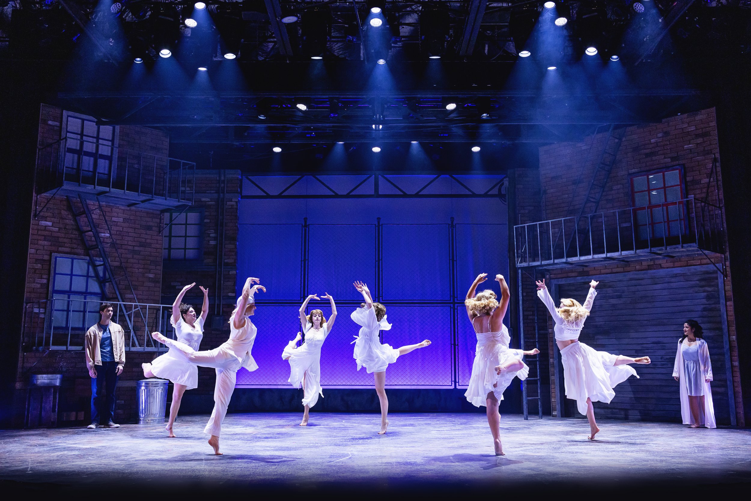 Theatre stage with eight dancers in white costumes performing ballet, flanked by two actors on each side and set designed to look like urban buildings.