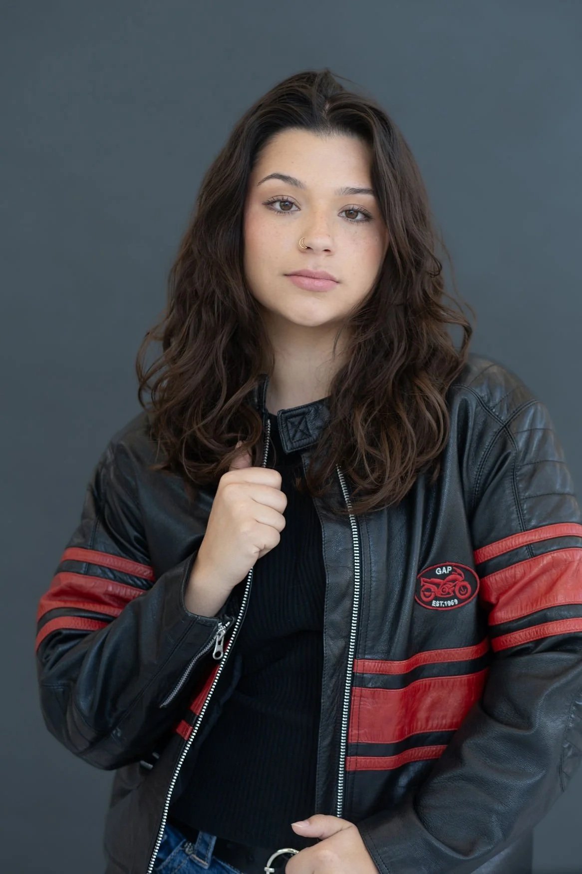 A young woman with long, wavy dark hair and light skin is posing against a plain gray background. She wears a black leather jacket with red stripes on the sleeves and a small logo on the chest, over a black top. She has a neutral expression, with her right hand holding the collar of her jacket and her left hand near her waist.