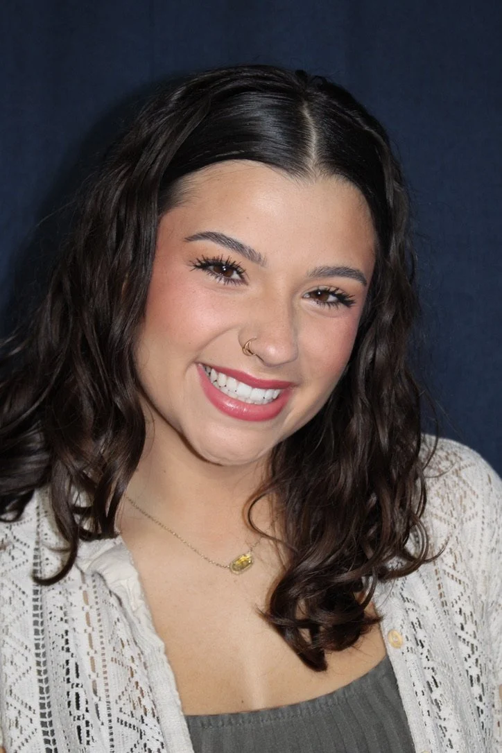 A young woman with long dark wavy hair, wearing a white crocheted top and a gold necklace, smiling at the camera against a dark background.
