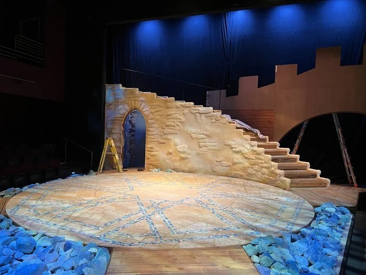 Stage set resembling a castle with stairs, a castle wall, and an arched doorway, surrounded by rocks, under blue lighting.