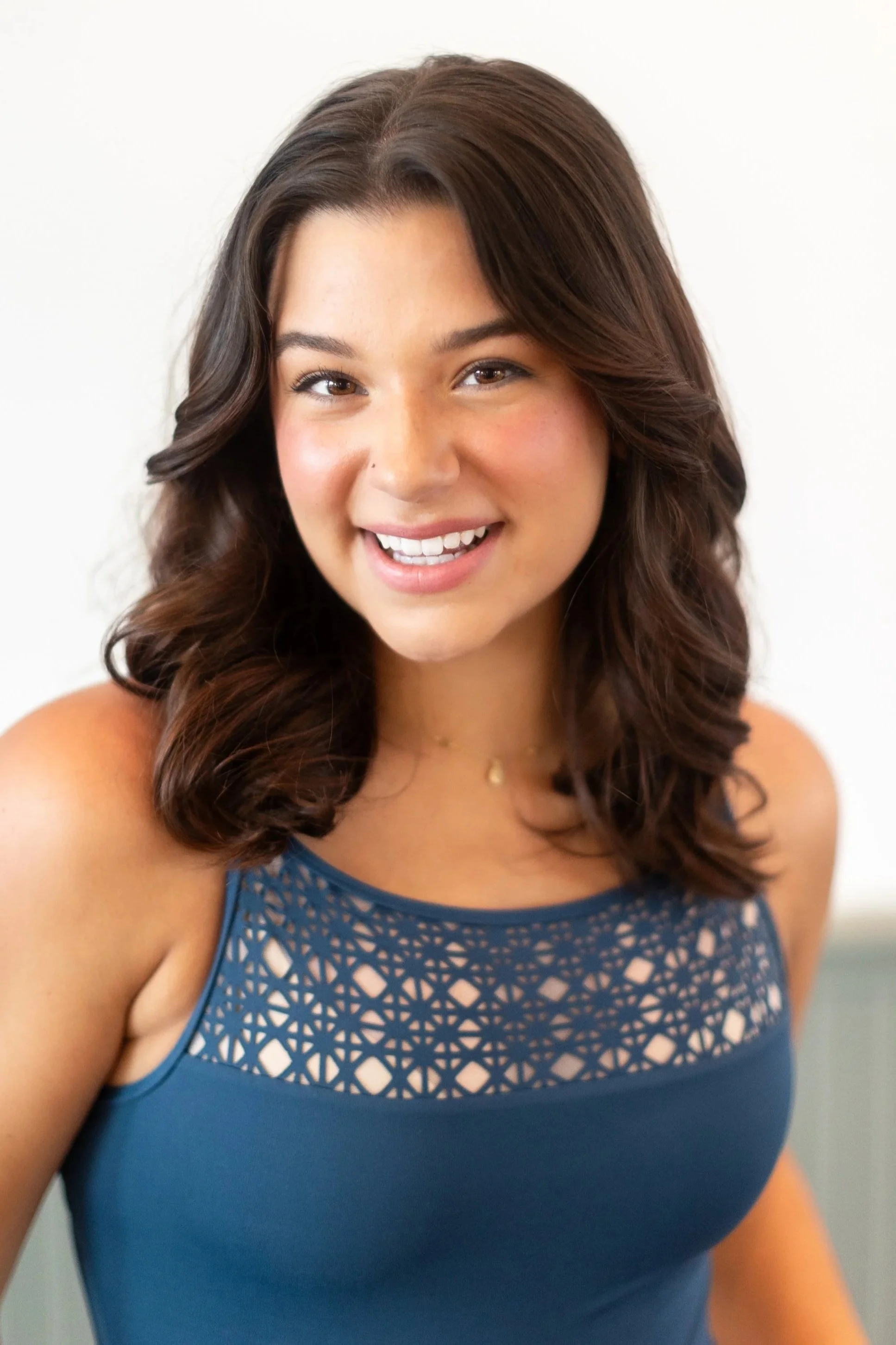 Young woman with wavy, shoulder-length brown hair smiling in a blue sleeveless top with a lace detail at the neckline.