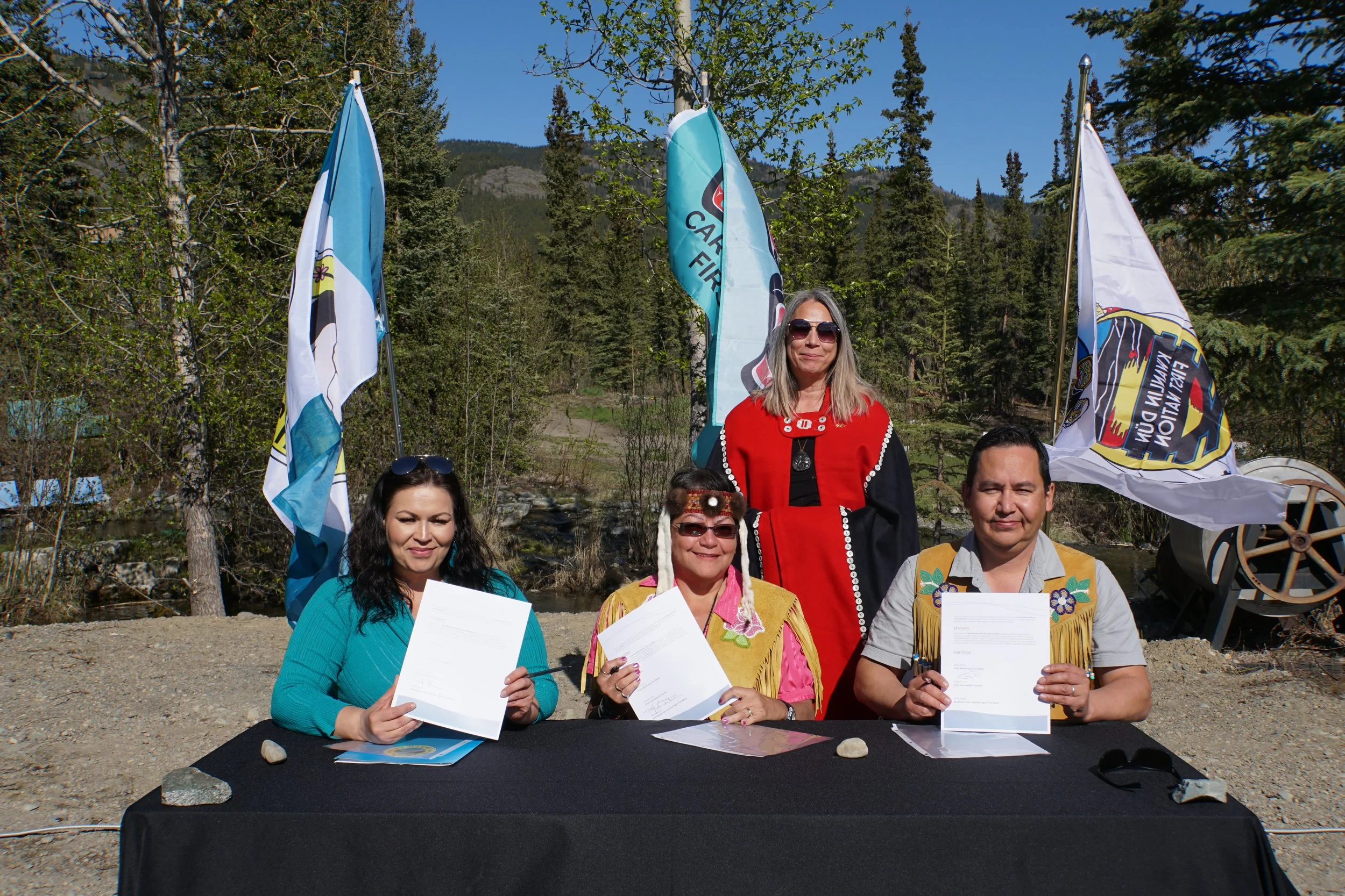 Four people sitting at a table outdoors, holding documents, with flags and trees in the background. One woman stands behind them, all smiling.