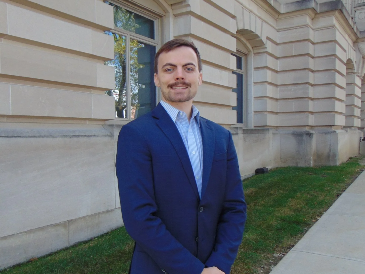 A young man with short brown hair, a light beard, and blue eyes is standing outdoors in front of a historic beige stone building. He is dressed in a dark blue suit jacket over a light blue striped shirt, smiling slightly with his hands clasped in front of him. Behind him, a large window and a sidewalk are visible, with some greenery and trees reflected in the window.