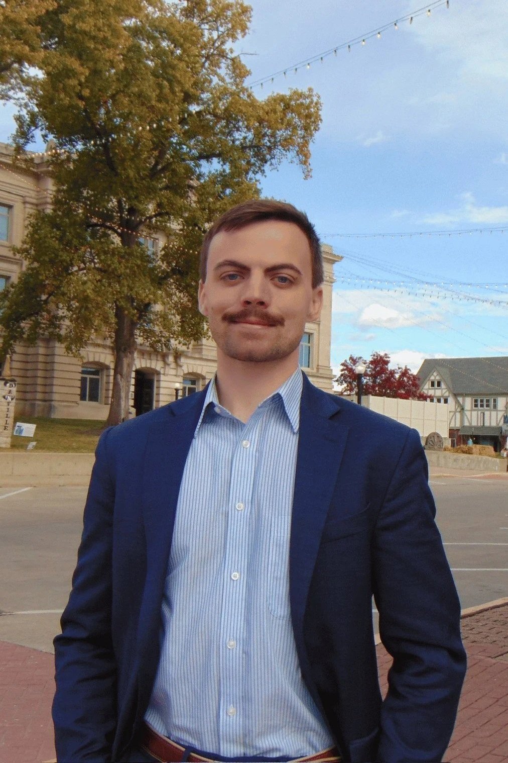 A man with a mustache and short dark hair dressed in a blue blazer and light blue striped shirt standing outdoors in front of trees and historic buildings under a clear sky with string lights above.