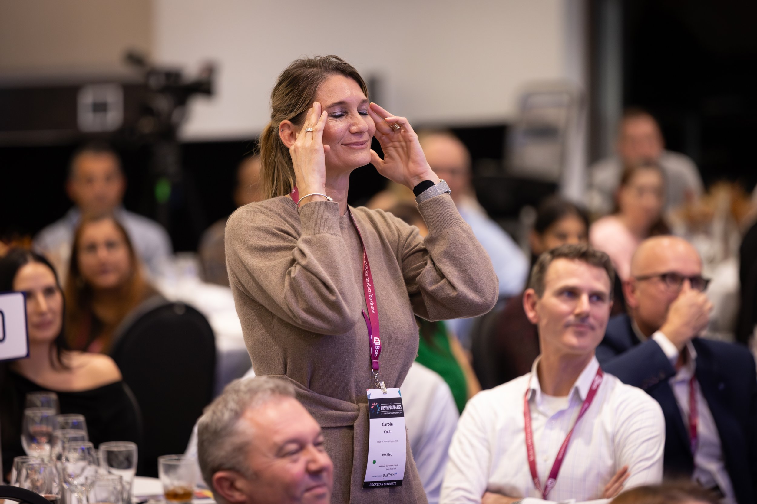 A woman standing with her hands on her temples, participating in a conference or seminar, surrounded by seated attendees.
