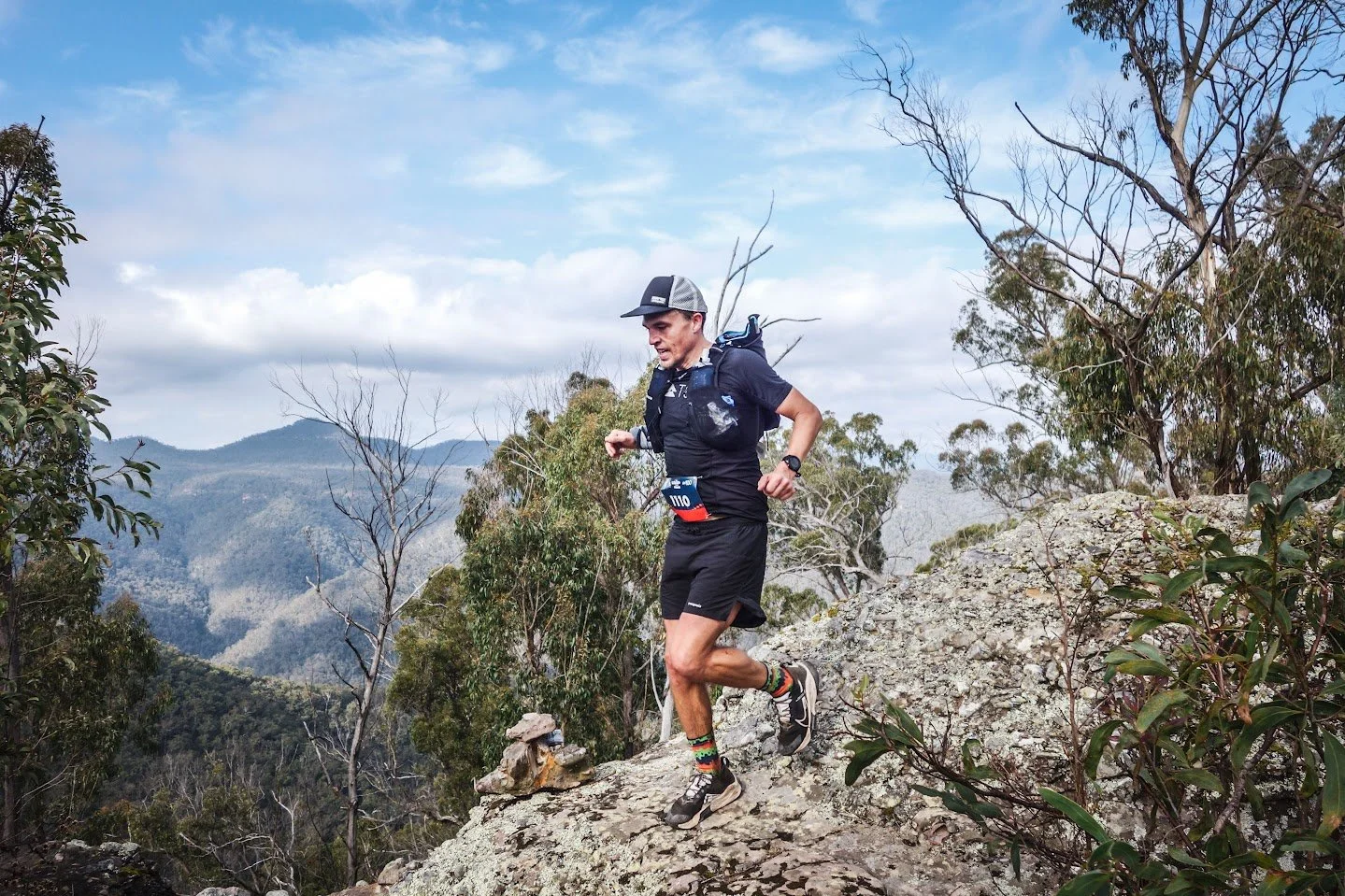 A man running on a rocky trail in a mountainous and forested area, wearing a black athletic outfit, a gray cap, a backpack, and colorful socks.